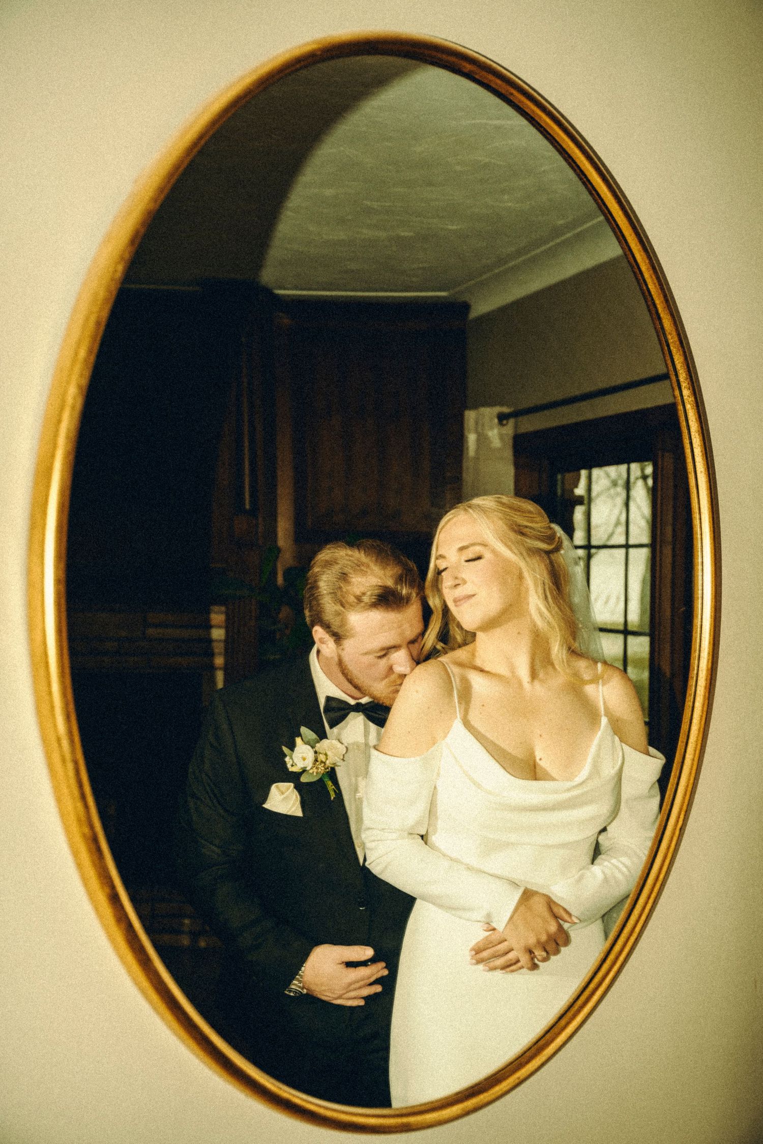 Bride and groom reflected in oval gold mirror, groom kissing bride's neck in romantic wedding portrait.