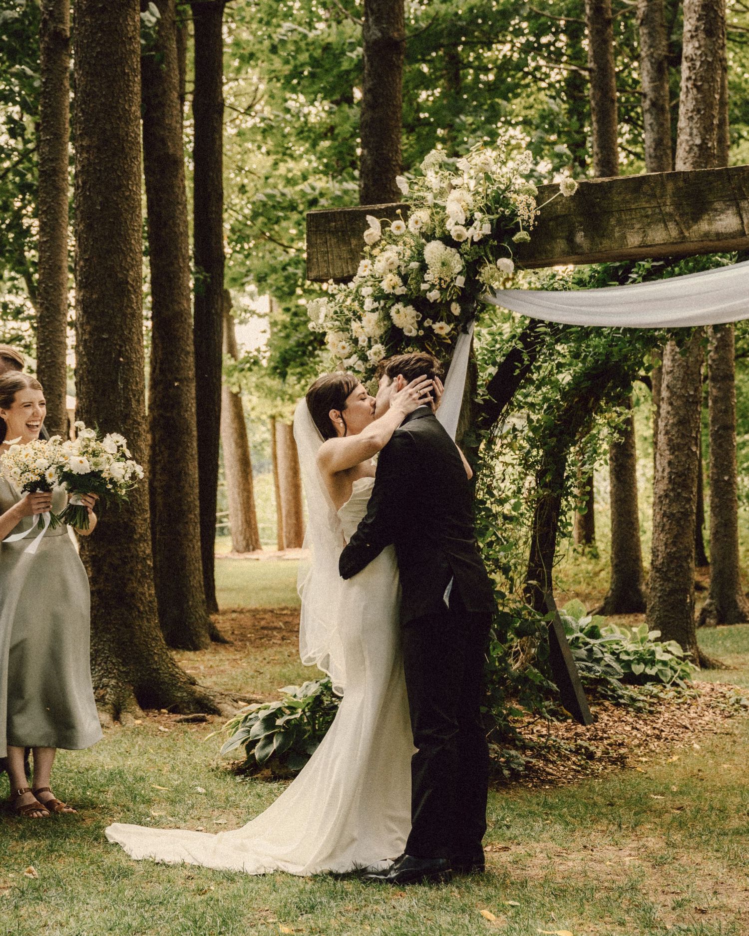 Bride and groom share first kiss under floral arch surrounded by tall trees at outdoor ceremony.