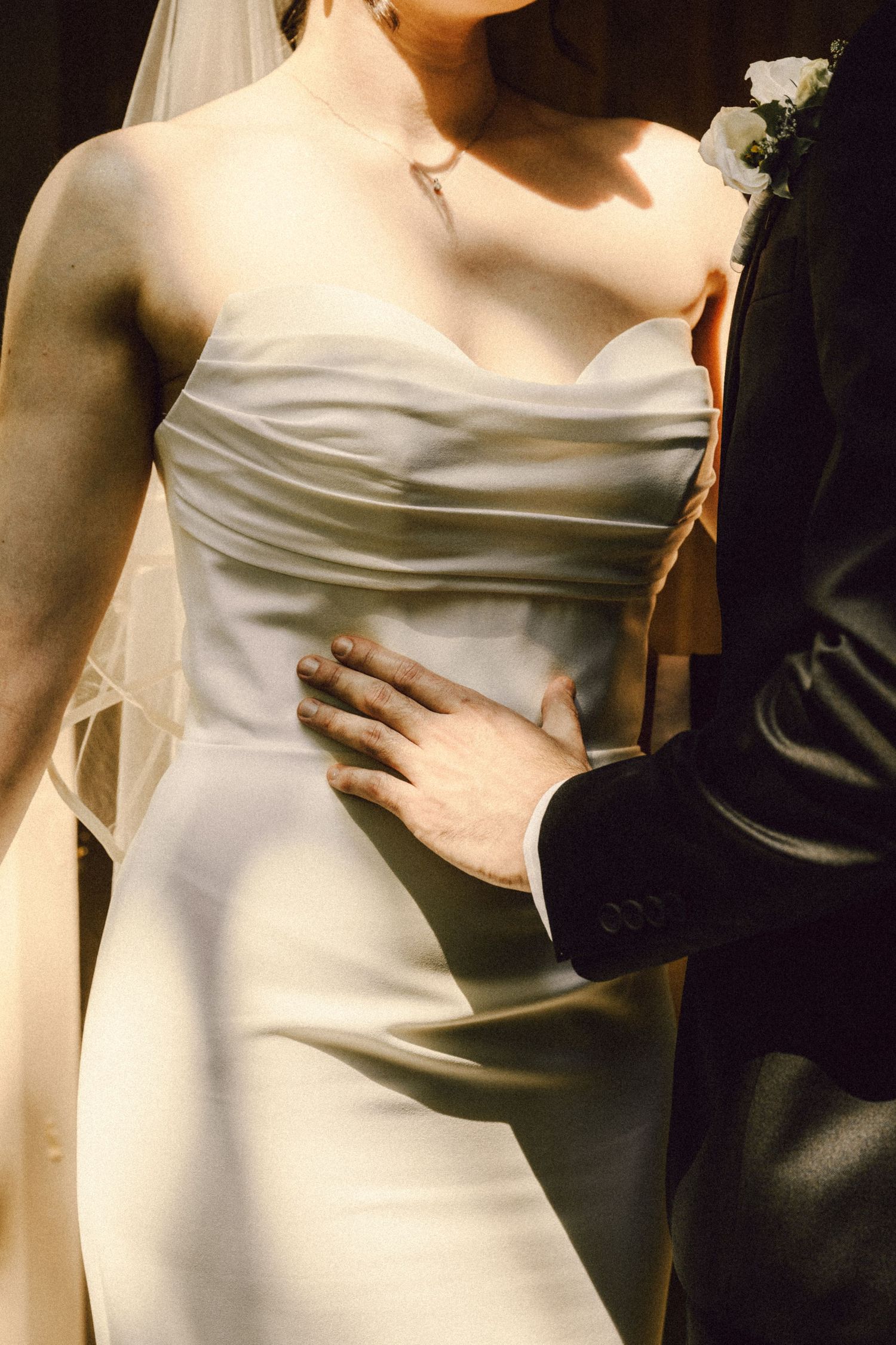 Groom's hand resting on bride's waist in elegant strapless white satin wedding gown with veil.