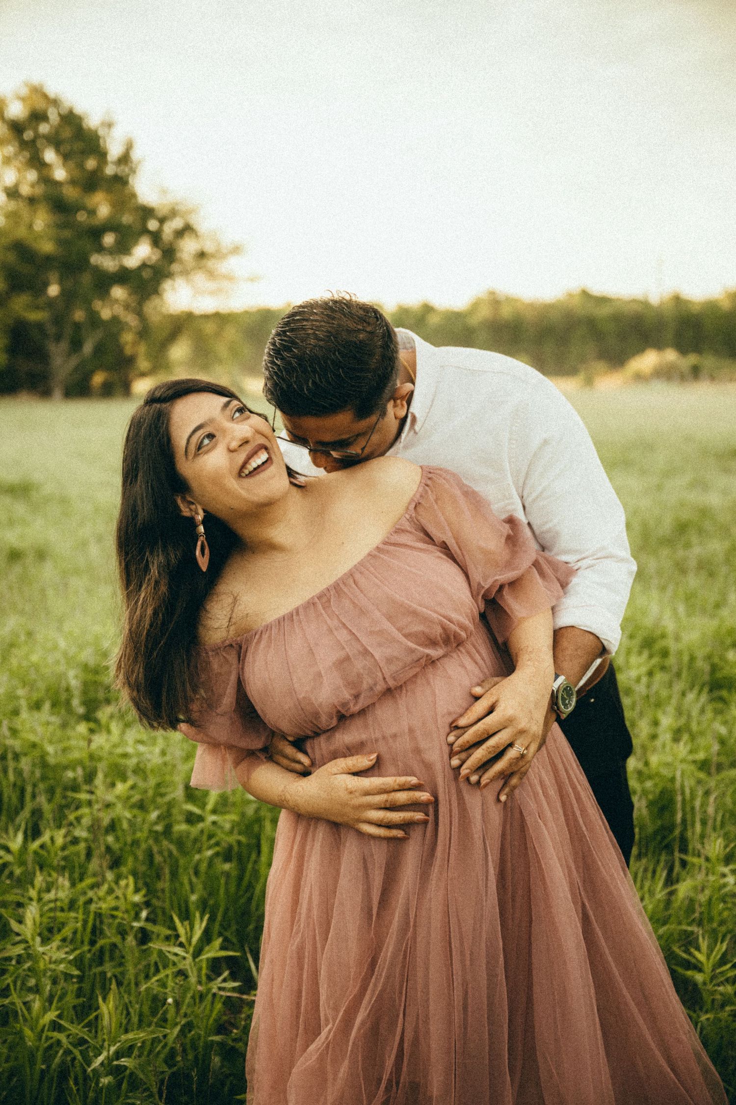 Pregnant woman in pink tulle gown laughing as partner embraces her in a lush green field.