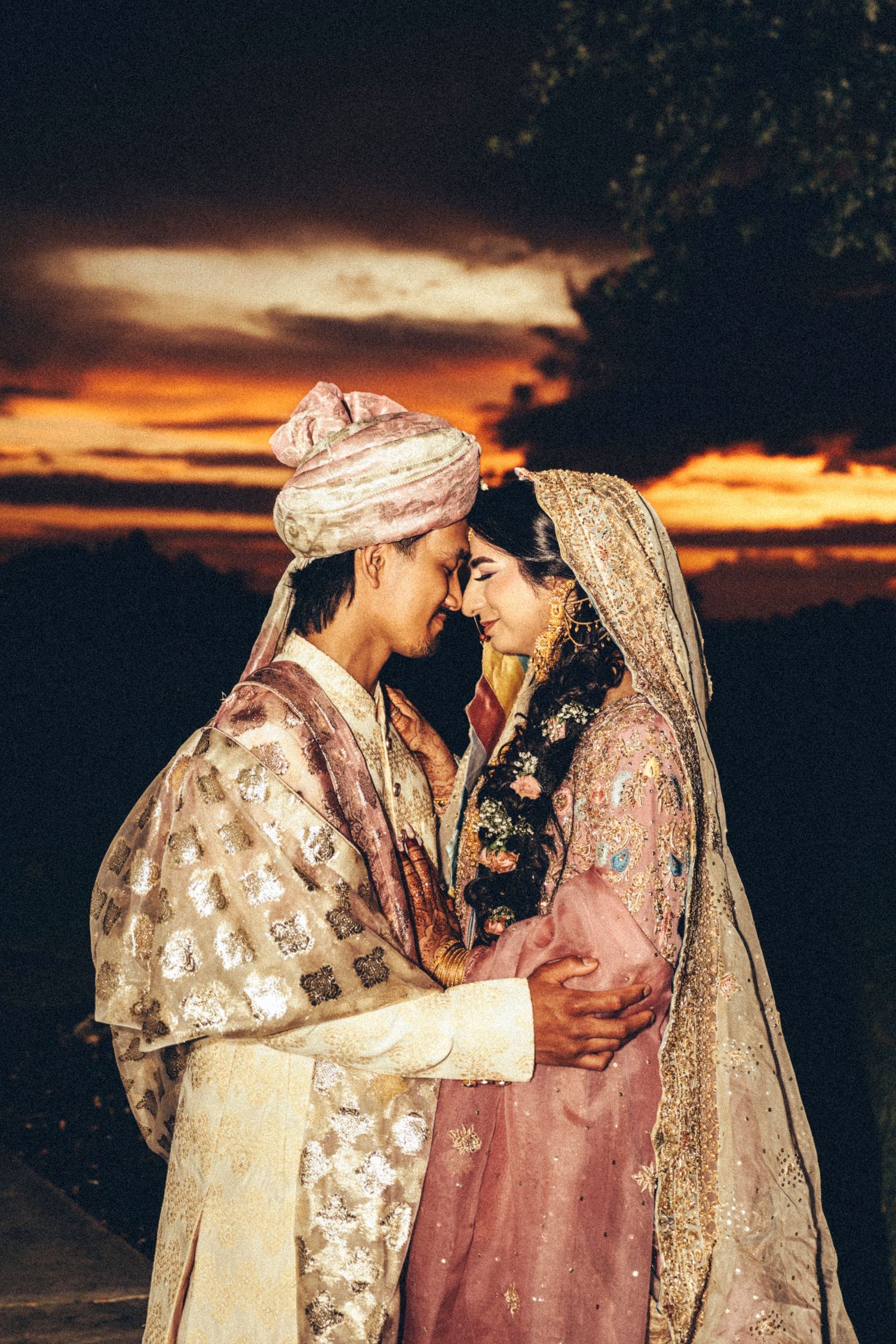South Asian bride and groom in traditional attire share an intimate forehead touch at sunset.
