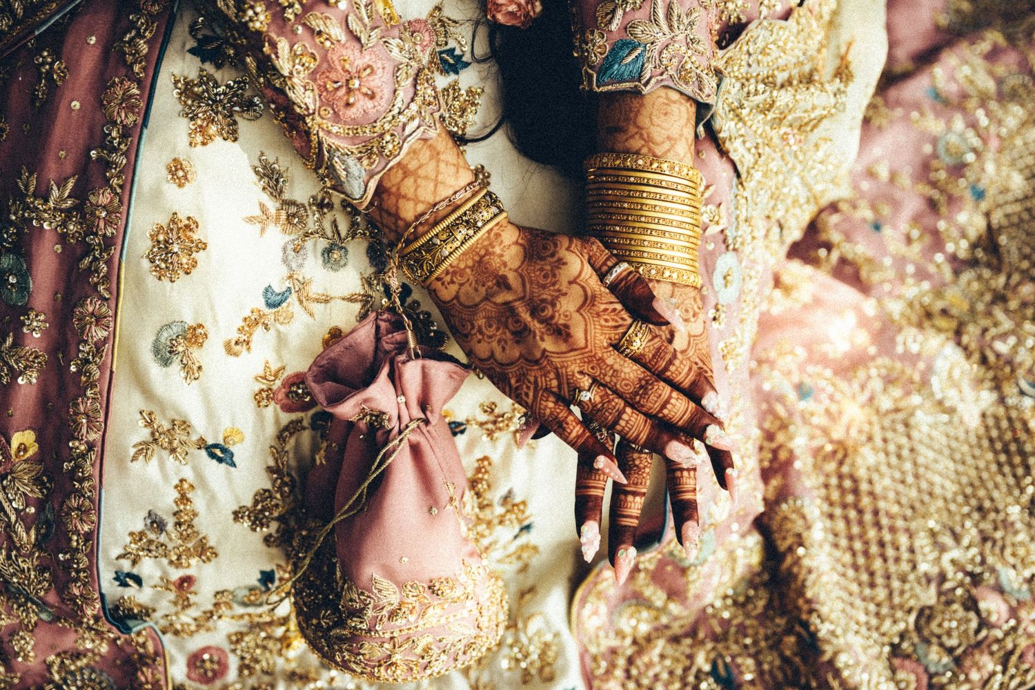 Close-up of South Asian bride's hands adorned with detailed henna mehndi, gold bangles, and embroidered pink lehenga.