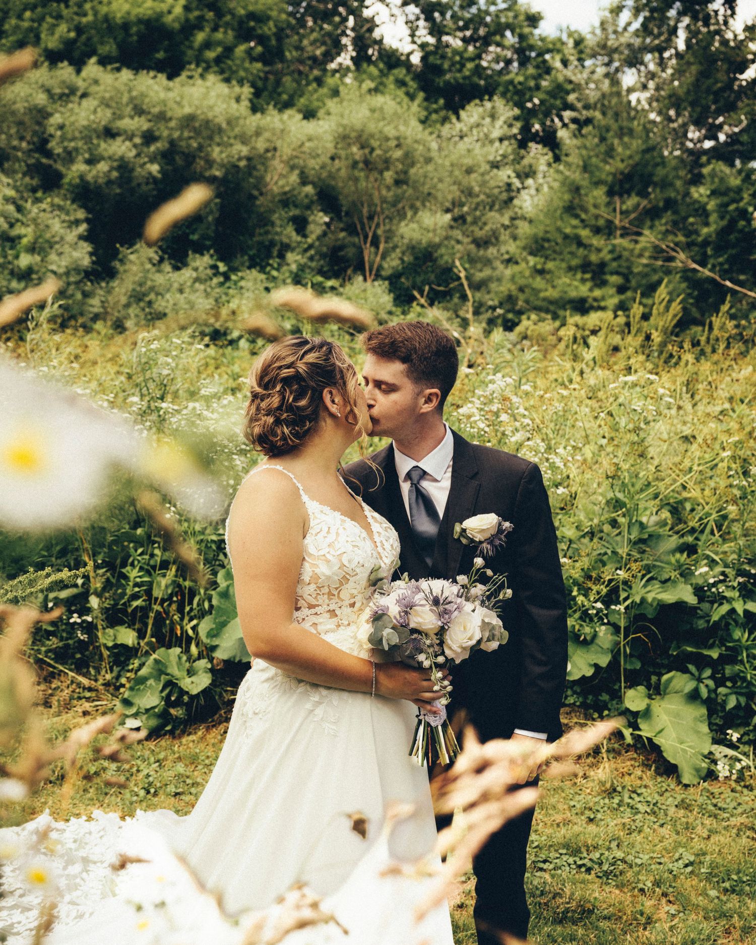Bride and groom share a romantic kiss surrounded by wildflowers and lush greenery outdoors.