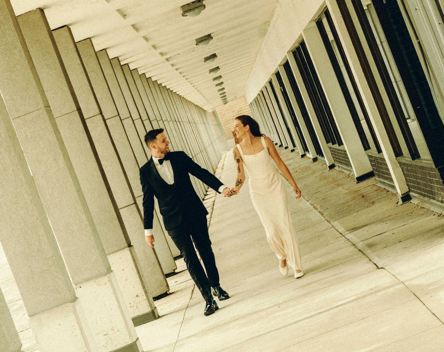 Bride and groom walking hand in hand through a modern architectural colonnade.