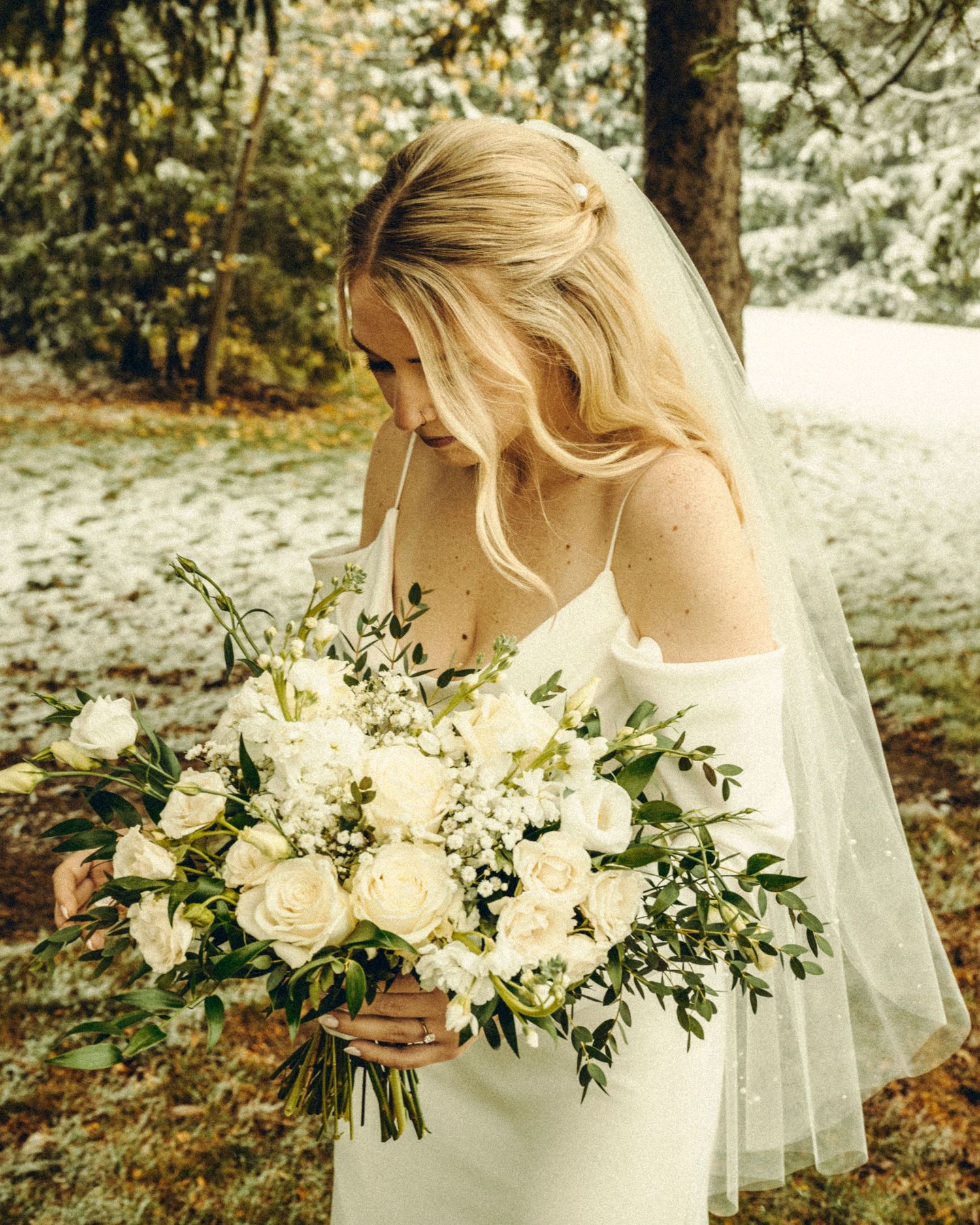 Blonde bride in white gown and veil looking down at lush white rose and greenery bouquet outdoors.