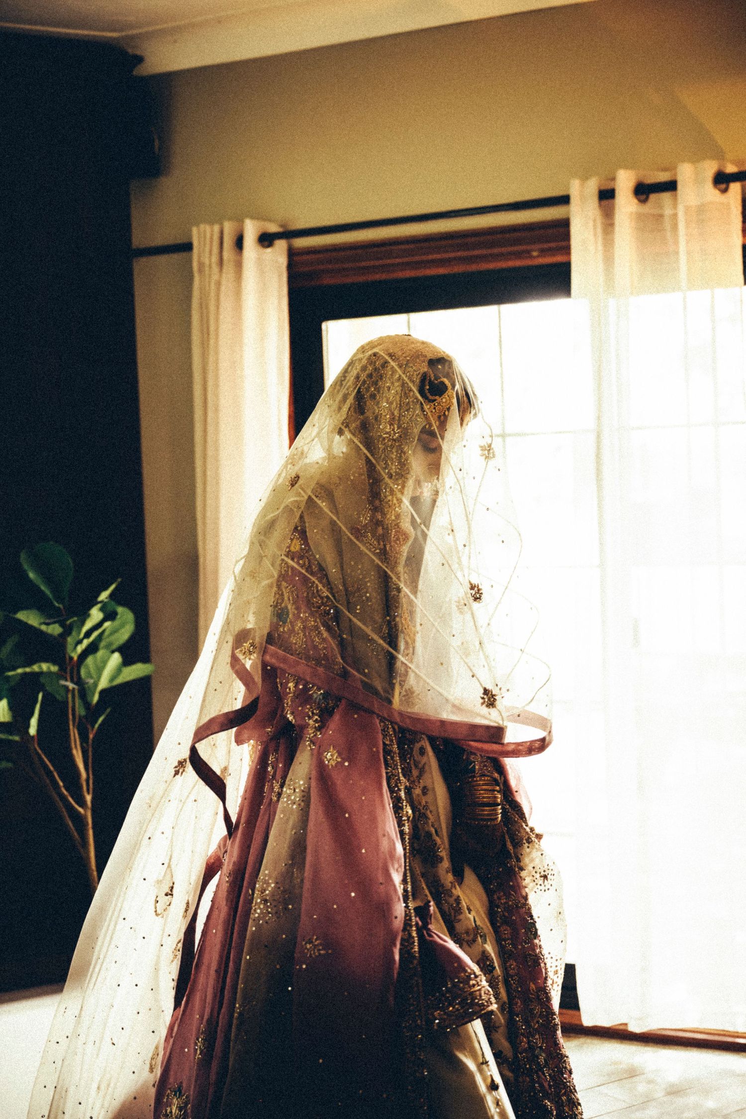 South Asian bride in a pink lehenga and embroidered veil stands backlit by a window in a serene bridal preparation moment.
