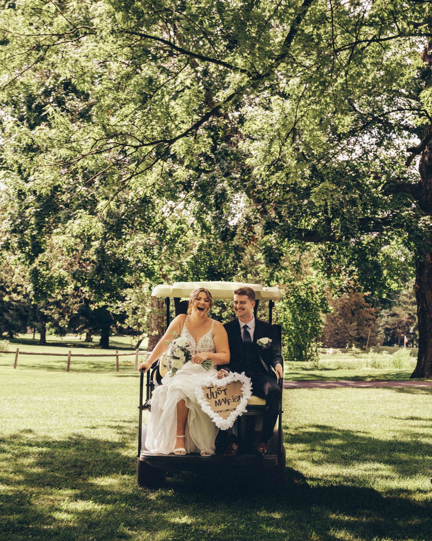 Smiling bride and groom ride a golf cart through lush green park grounds on wedding day.