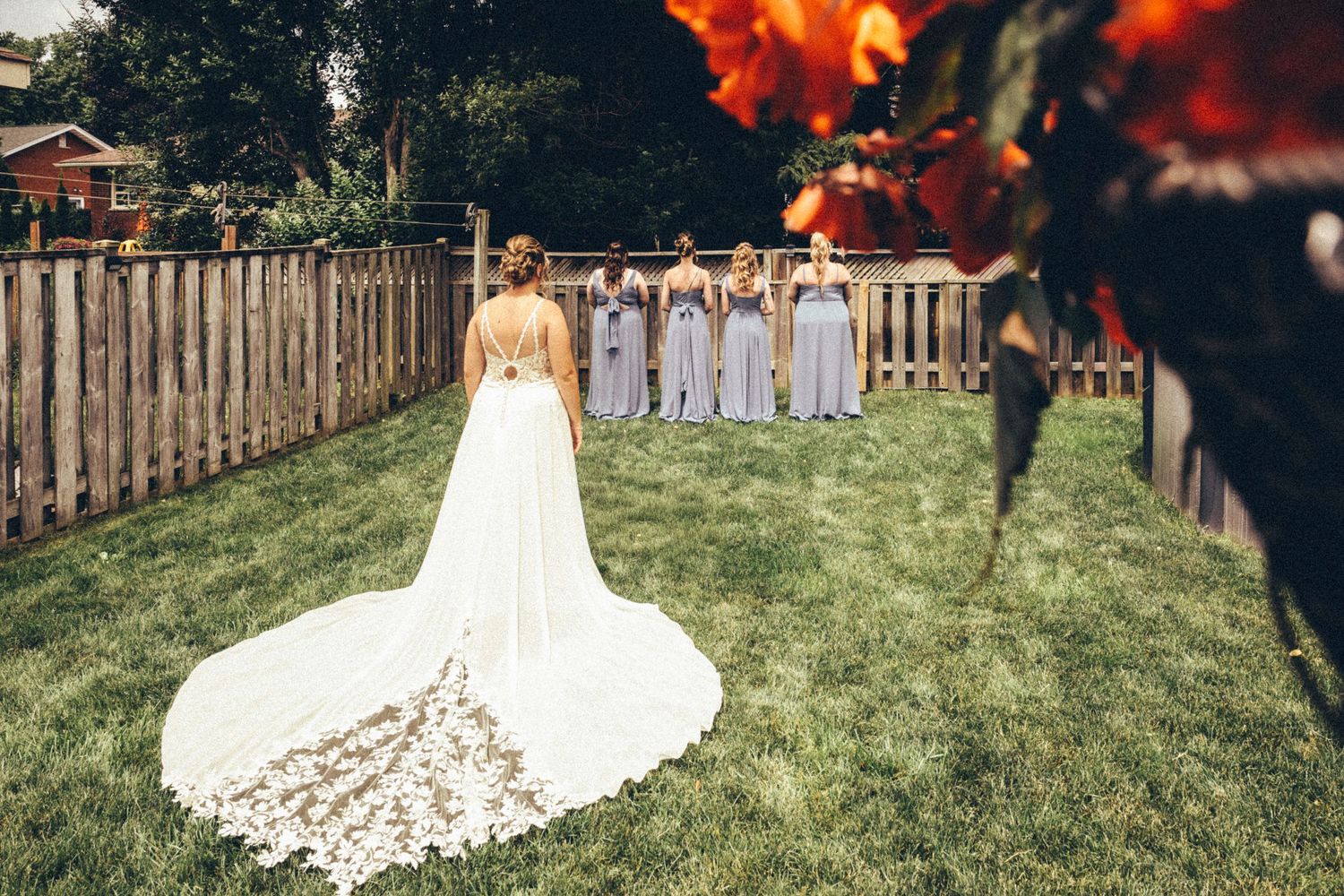 Bride in lace-trained wedding gown facing bridesmaids in gray dresses across green lawn.