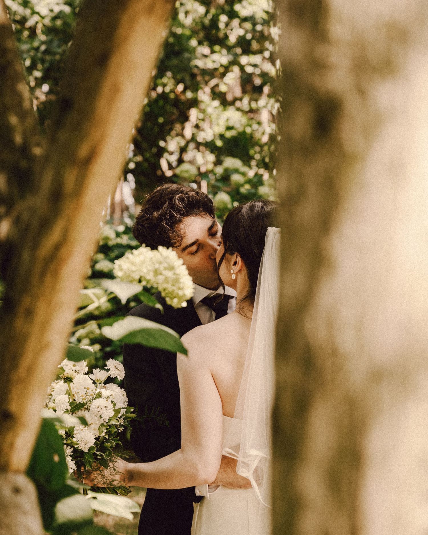 Newlyweds share a romantic kiss framed by tree trunks with white hydrangeas and a bridal bouquet visible.