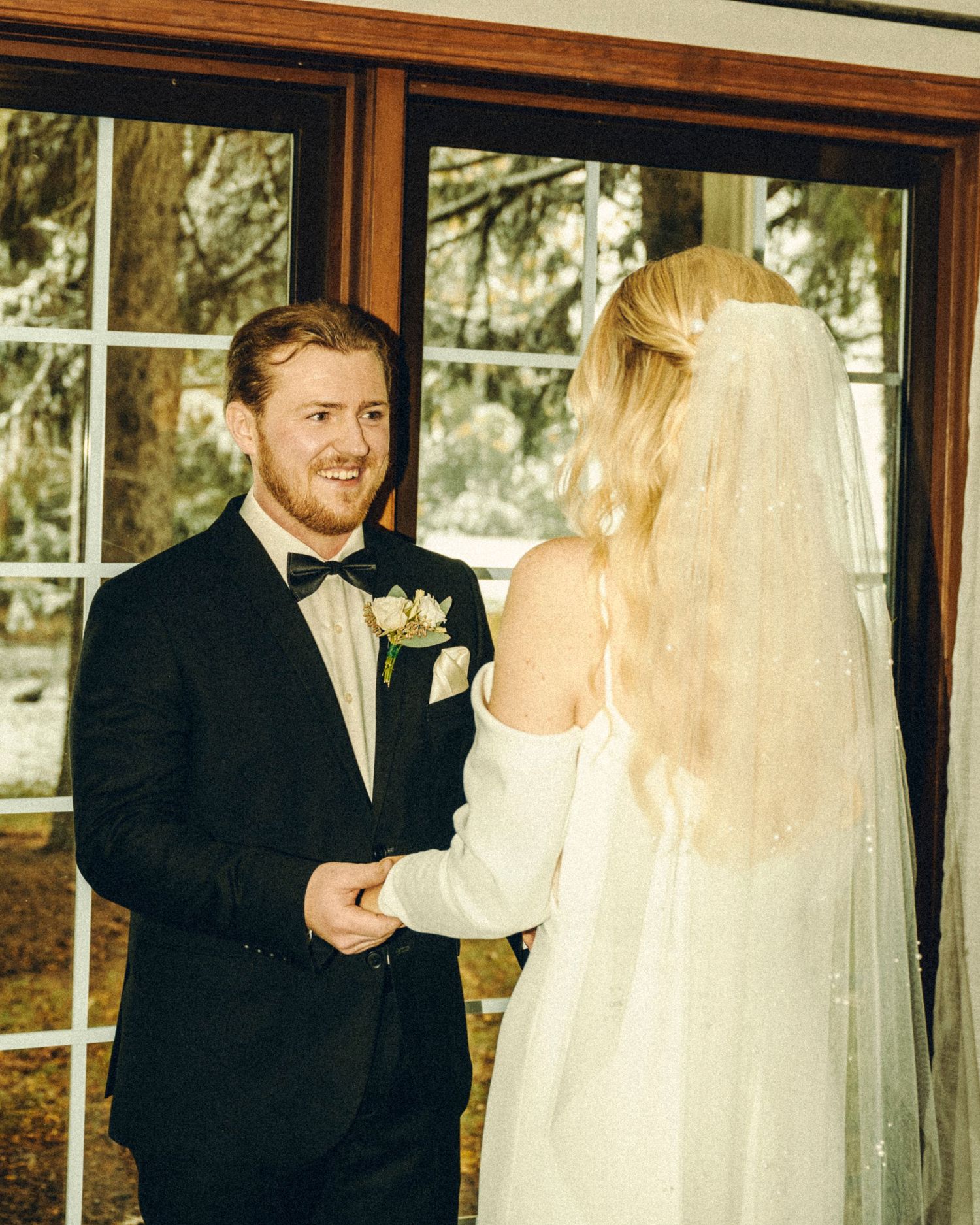 Groom in black tuxedo smiling at blonde bride in white off-shoulder gown during indoor wedding ceremony.