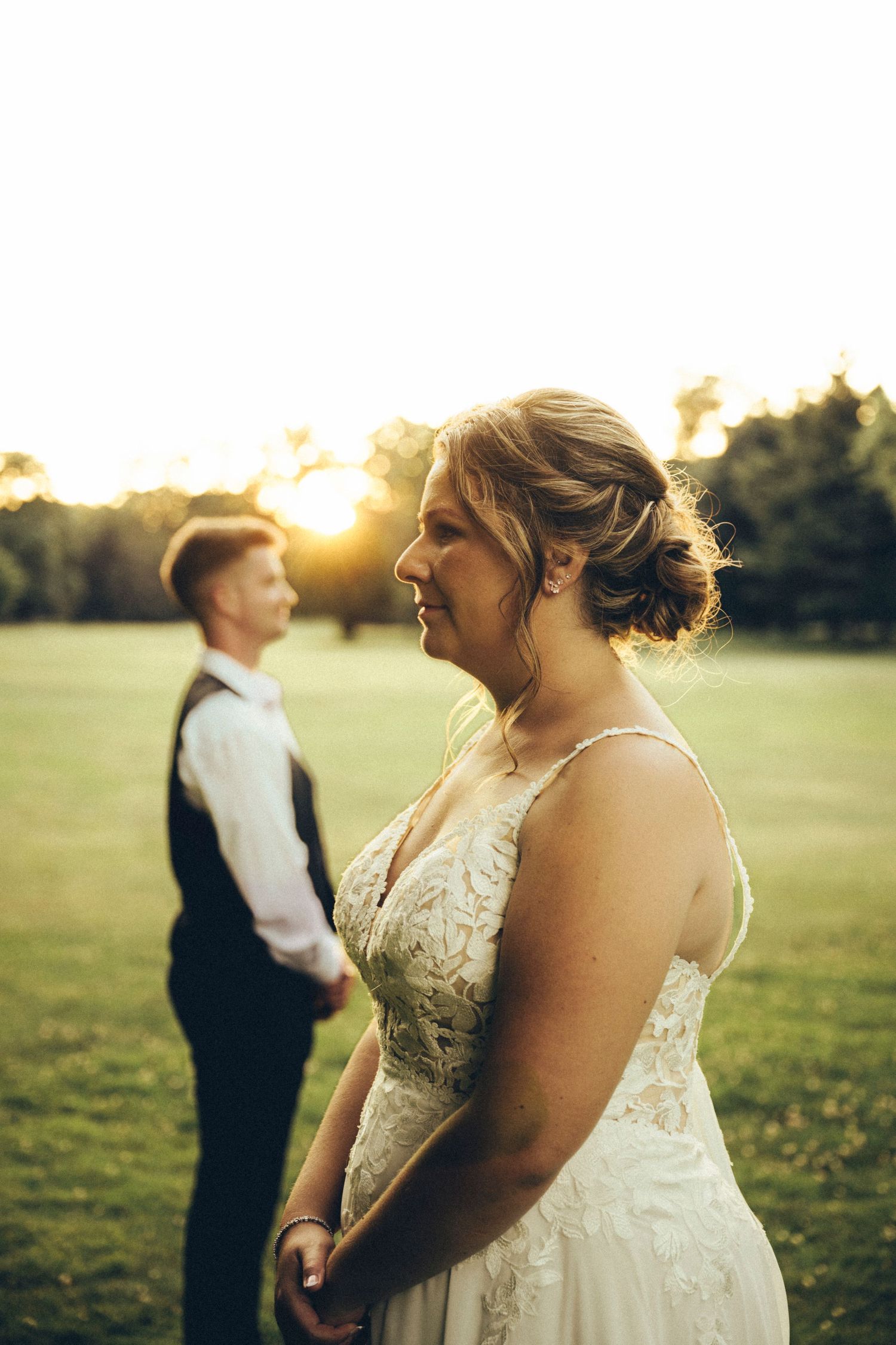 Smiling bride in lace wedding dress stands in foreground with groom at golden sunset.