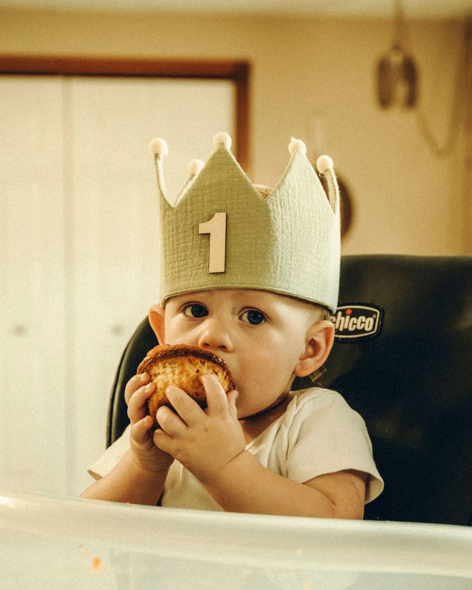 Baby wearing number 1 crown hat eating a muffin in a Chicco high chair.