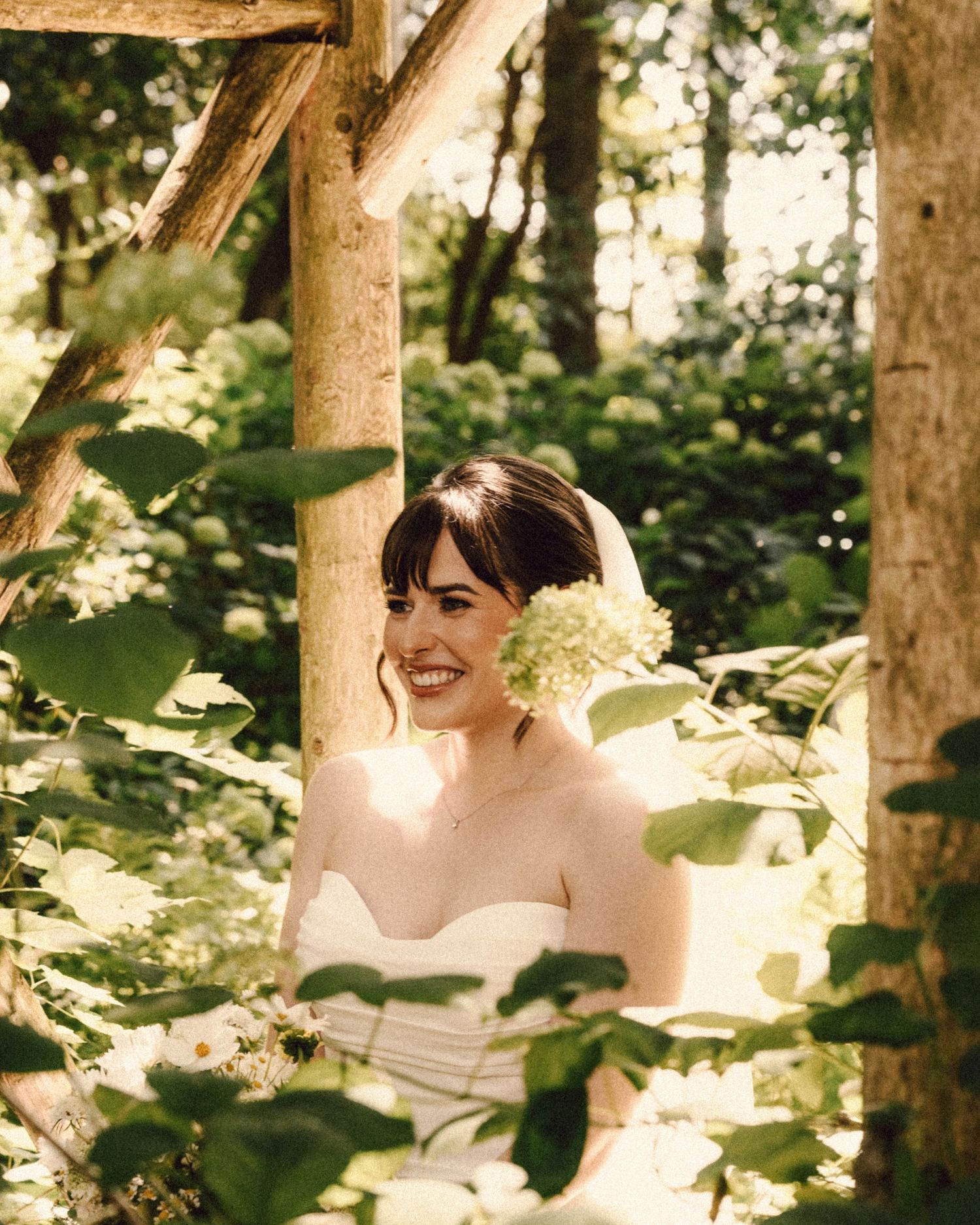 Radiant bride in strapless white gown smiles joyfully surrounded by lush greenery and white hydrangeas.