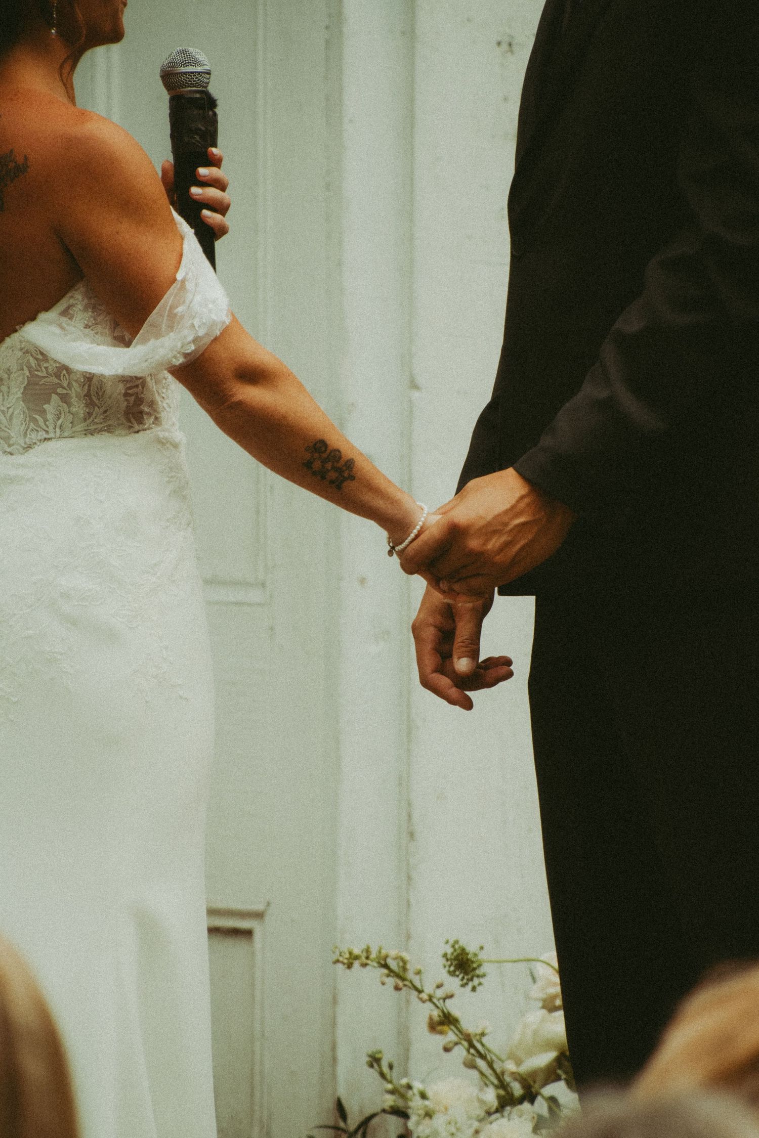Close-up of bride and groom holding hands during wedding ceremony, bride with floral arm tattoo.