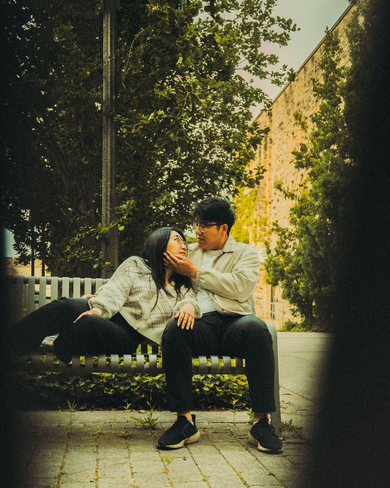 Couple sharing an intimate moment on a bench surrounded by green trees in an urban park.