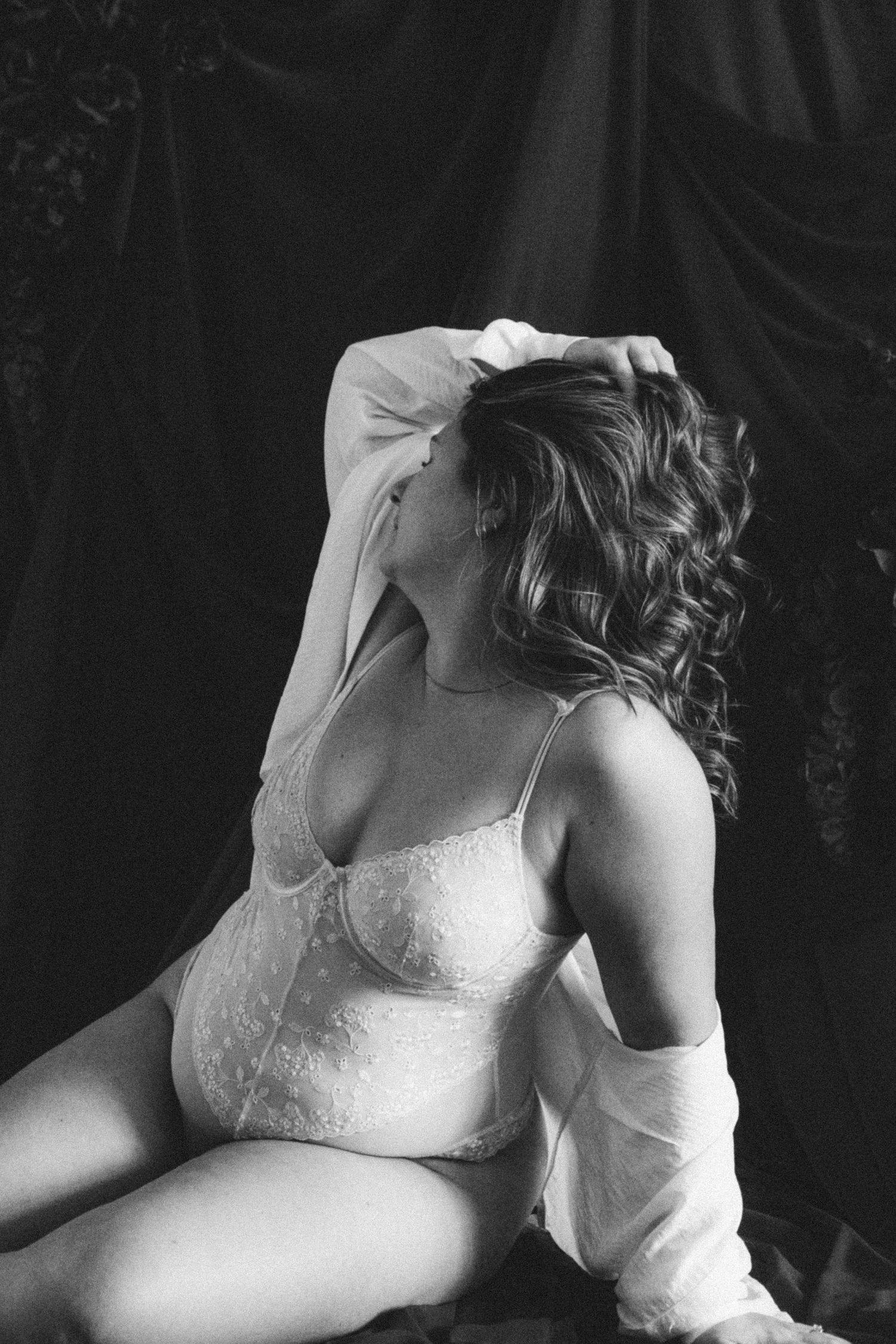 Black and white boudoir photo of woman in white lace lingerie seated gracefully against dramatic dark fabric backdrop.
