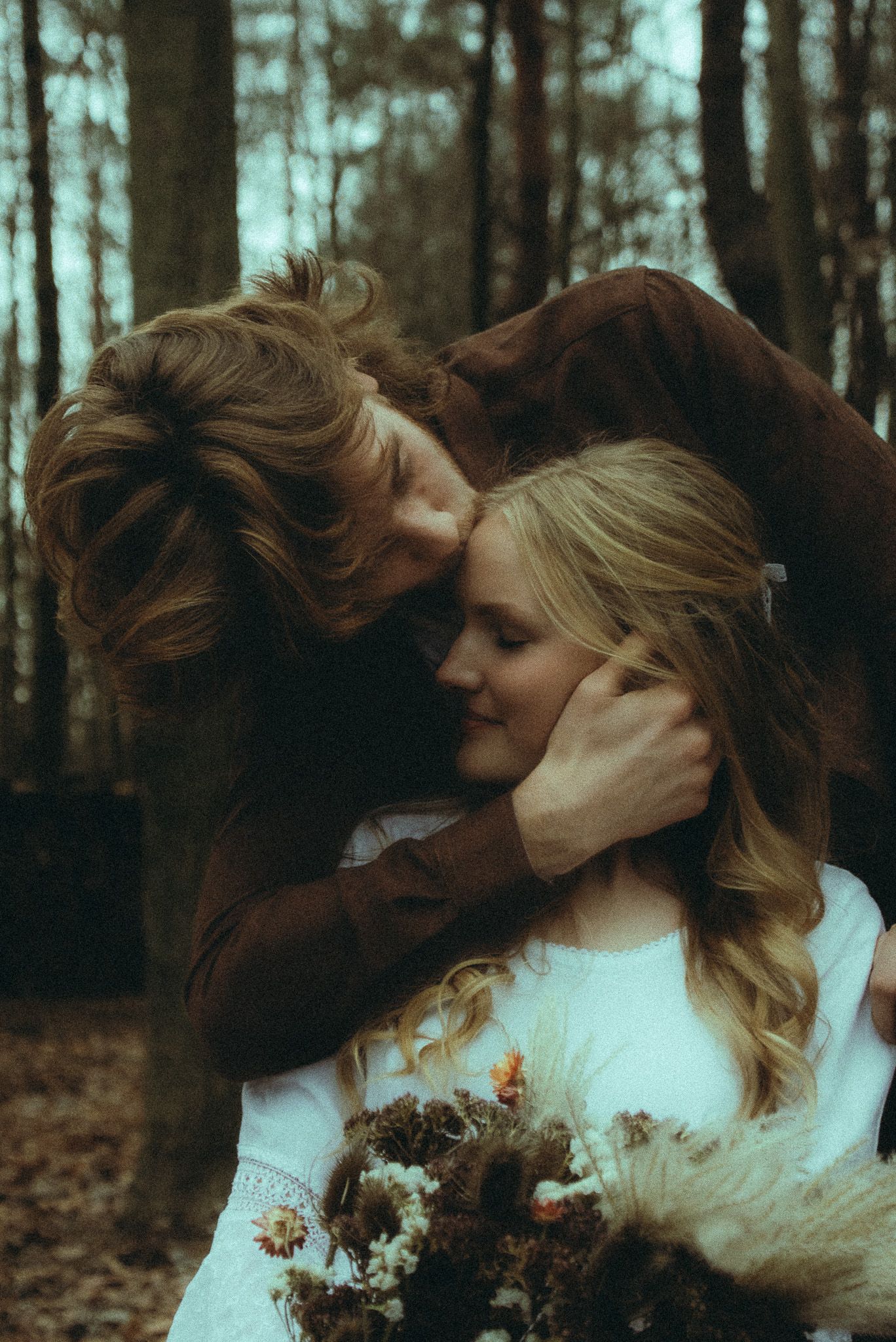 Man tenderly embracing woman holding a wildflower bouquet in a moody autumn forest setting.