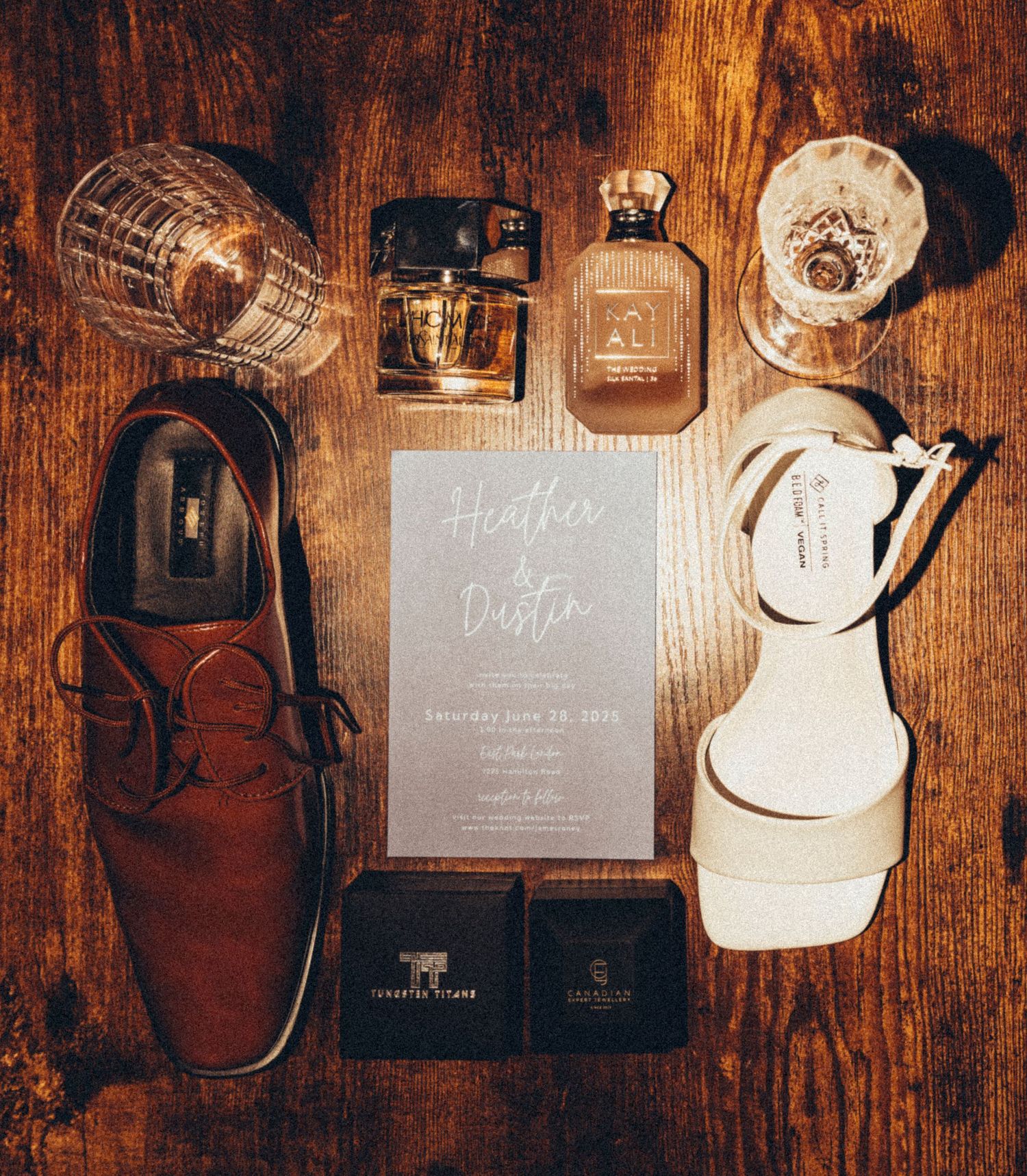 Wedding flat lay featuring invitation, brown loafers, white heels, perfume, whiskey glasses on wood.