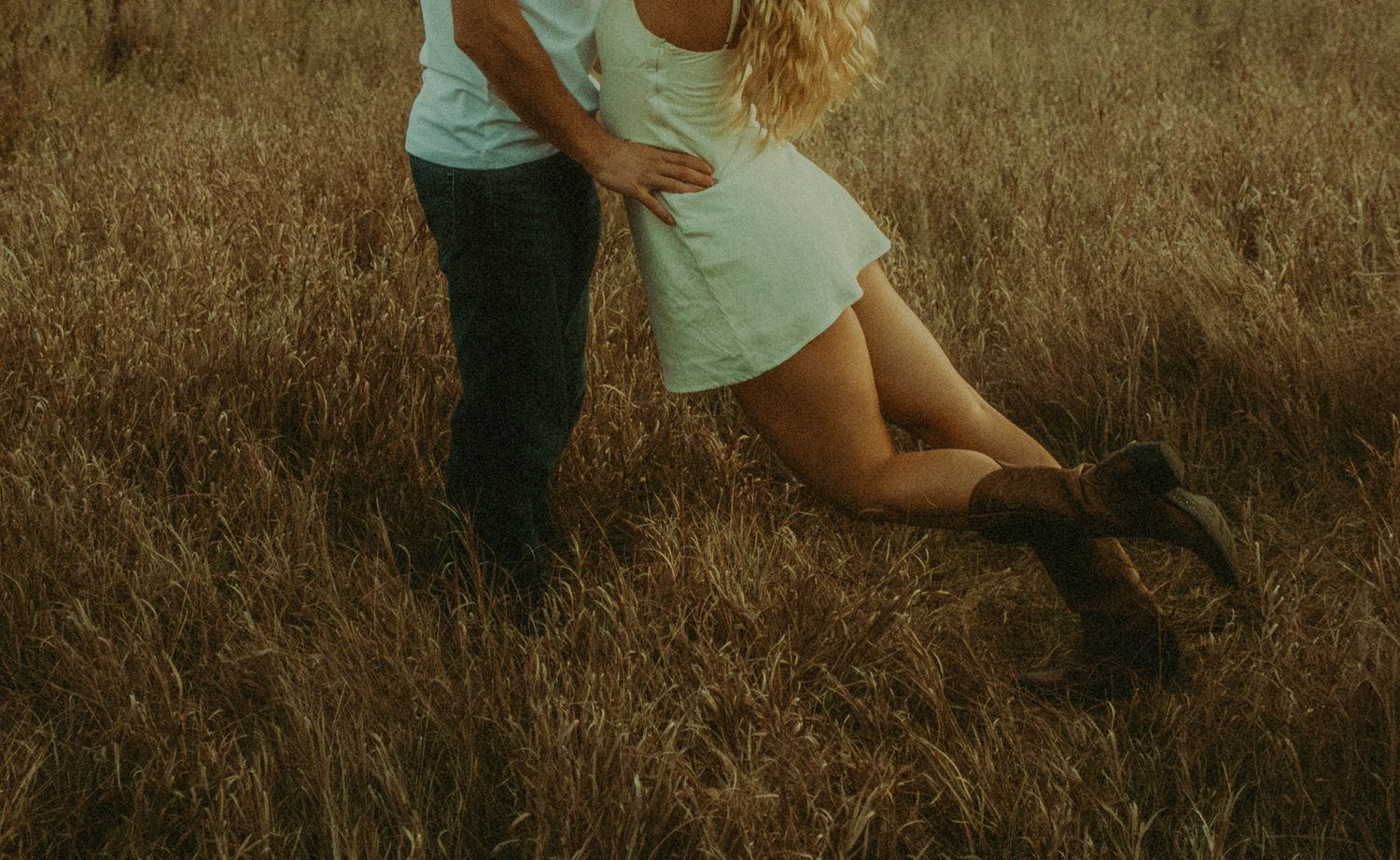Couple embracing romantically in a golden field of tall grass, woman wearing white dress and brown boots.