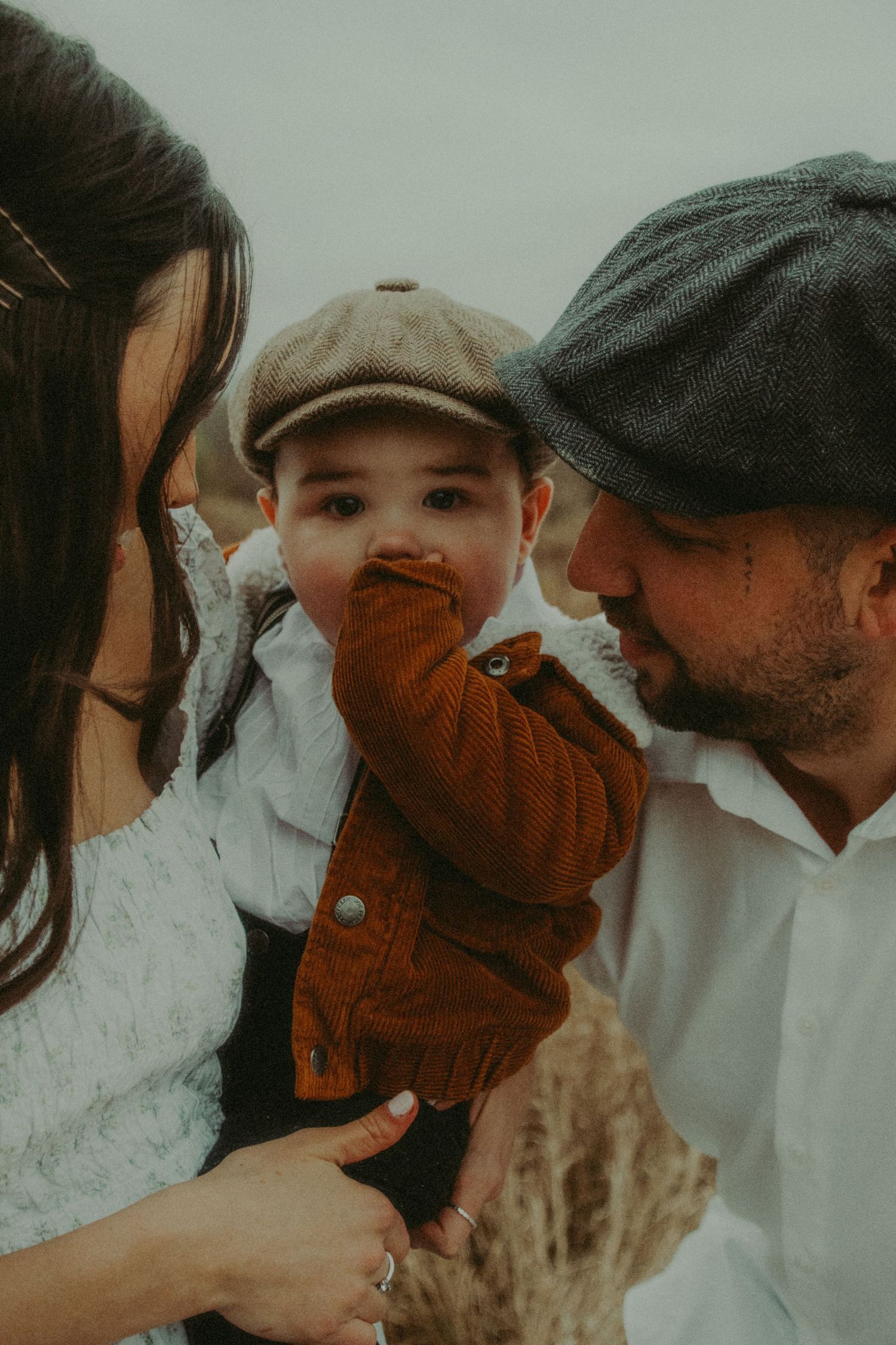 Parents lovingly kissing their baby wearing a rust orange jacket and newsboy cap outdoors.