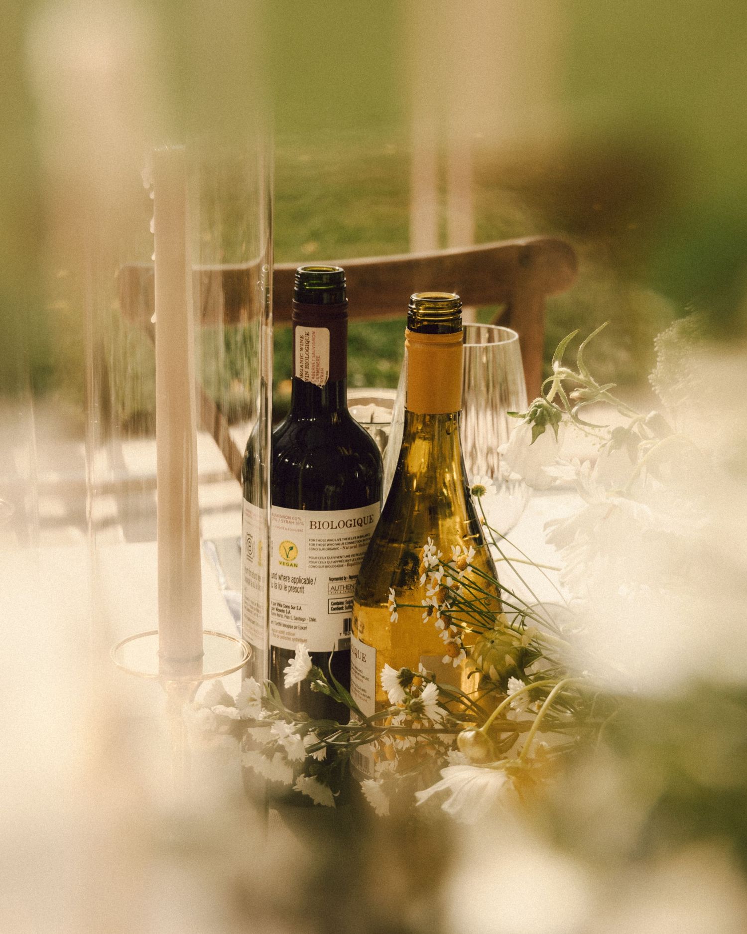 Two wine bottles, red and white, displayed among floral greenery on elegant outdoor wedding reception table.