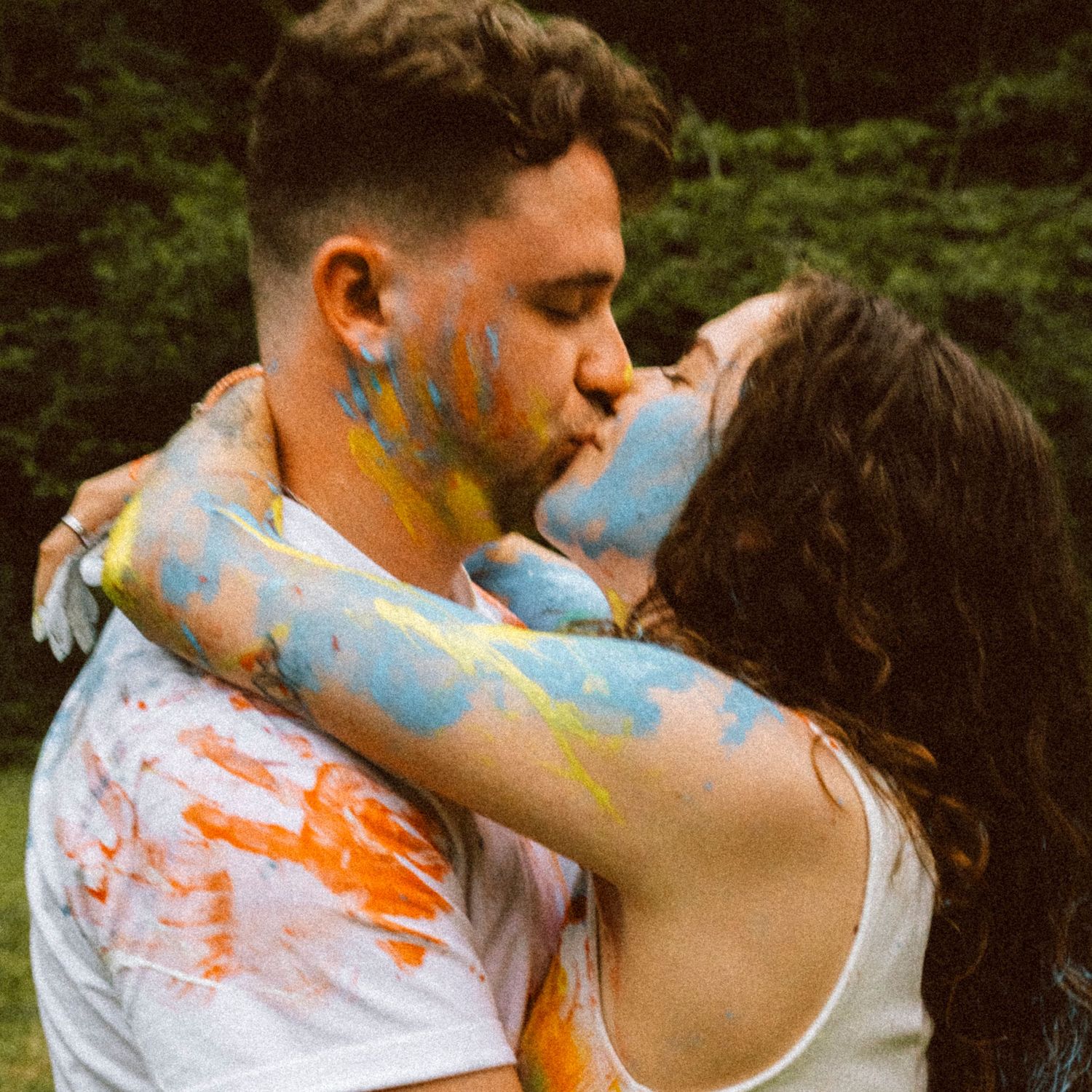 Couple kissing passionately while covered in colorful paint splatters outdoors in nature.