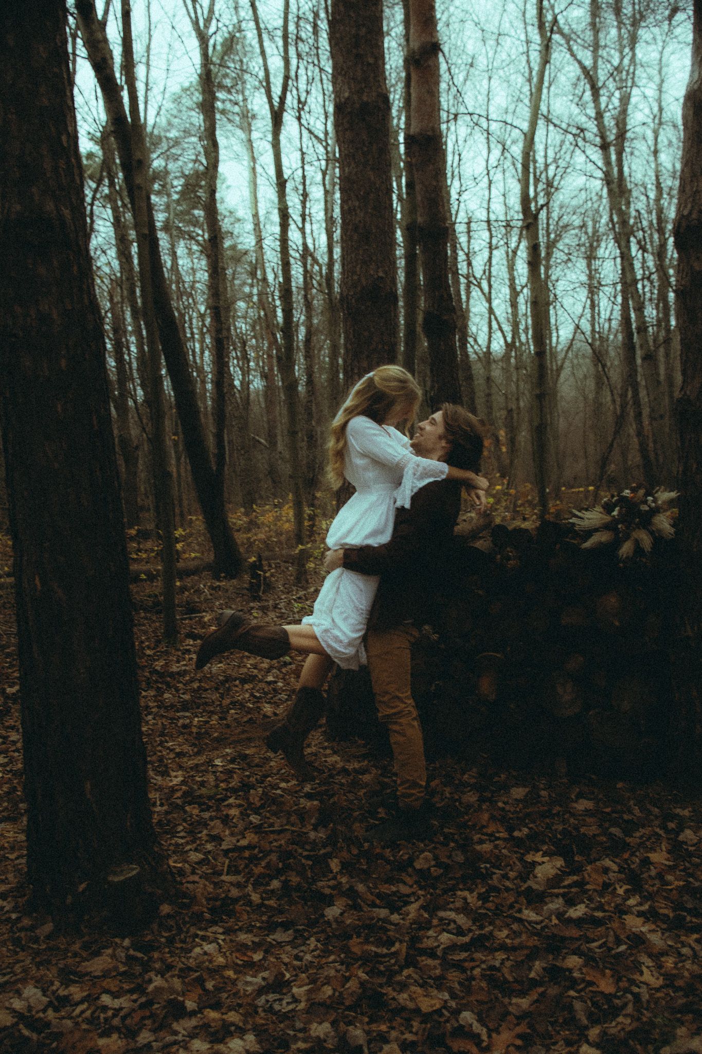 Couple embracing in a misty autumn forest with fallen leaves, man lifting woman in white dress.