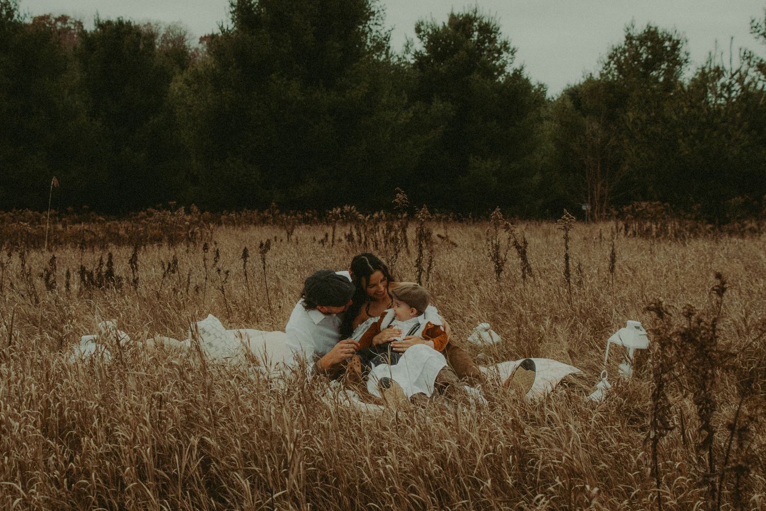 Family sitting together on white blanket in golden autumn field surrounded by tall grasses.
