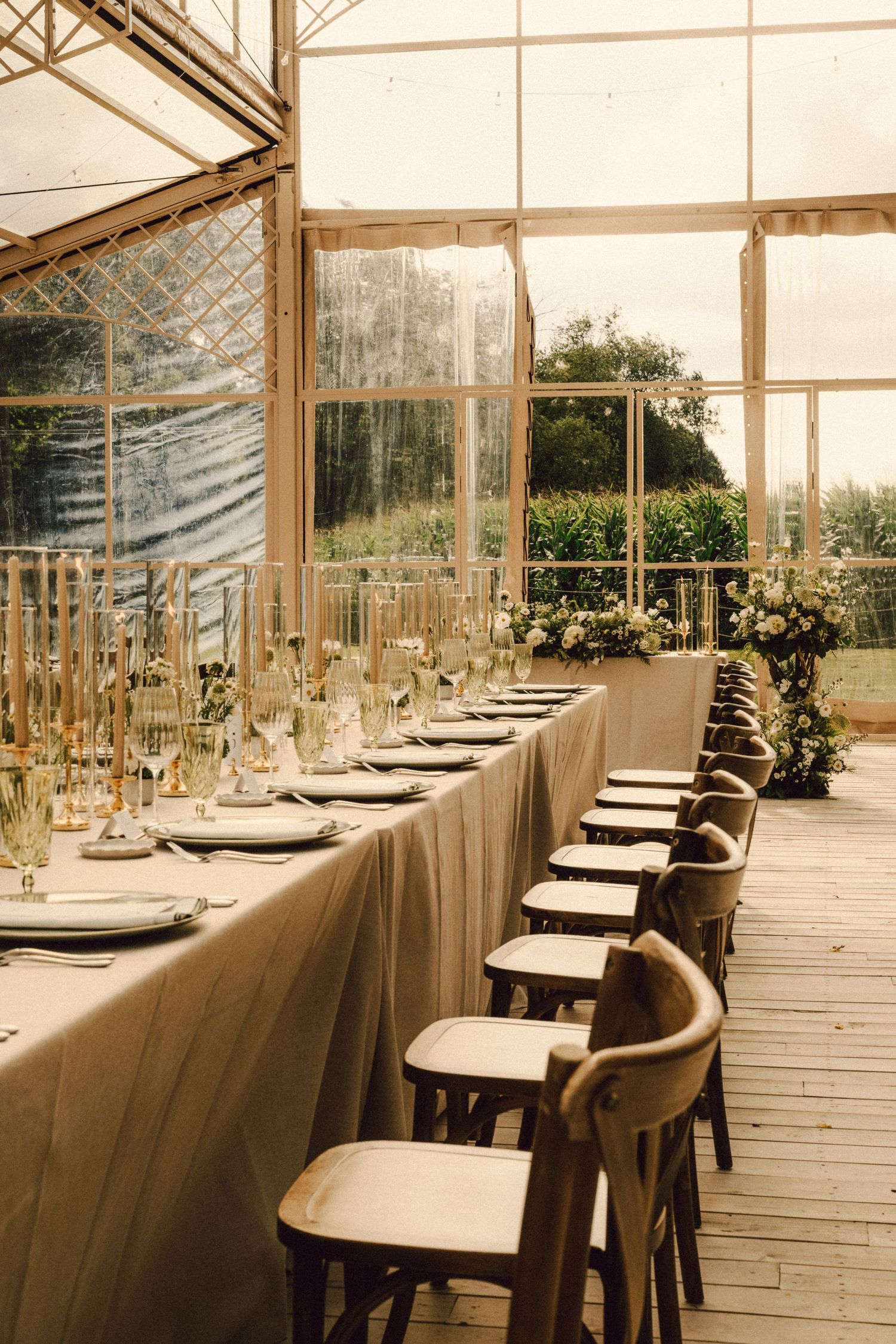 Elegant greenhouse wedding reception with long linen-draped tables, wooden chairs, gold candles and floral arrangements.