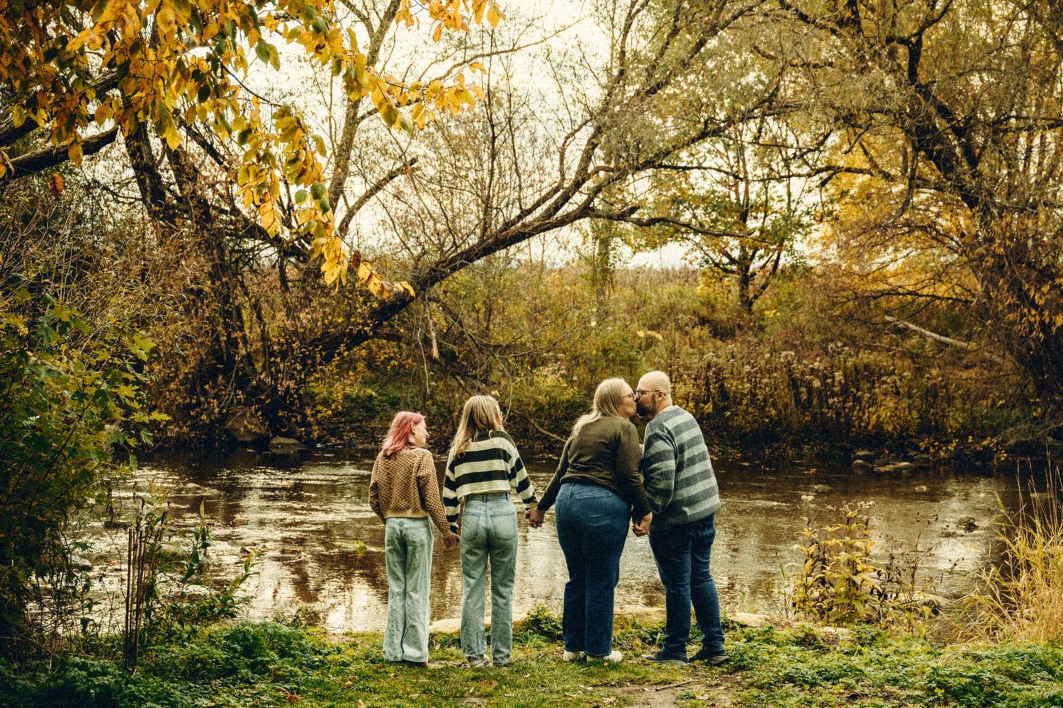 Family of four standing together viewing a peaceful pond surrounded by autumn foliage.
