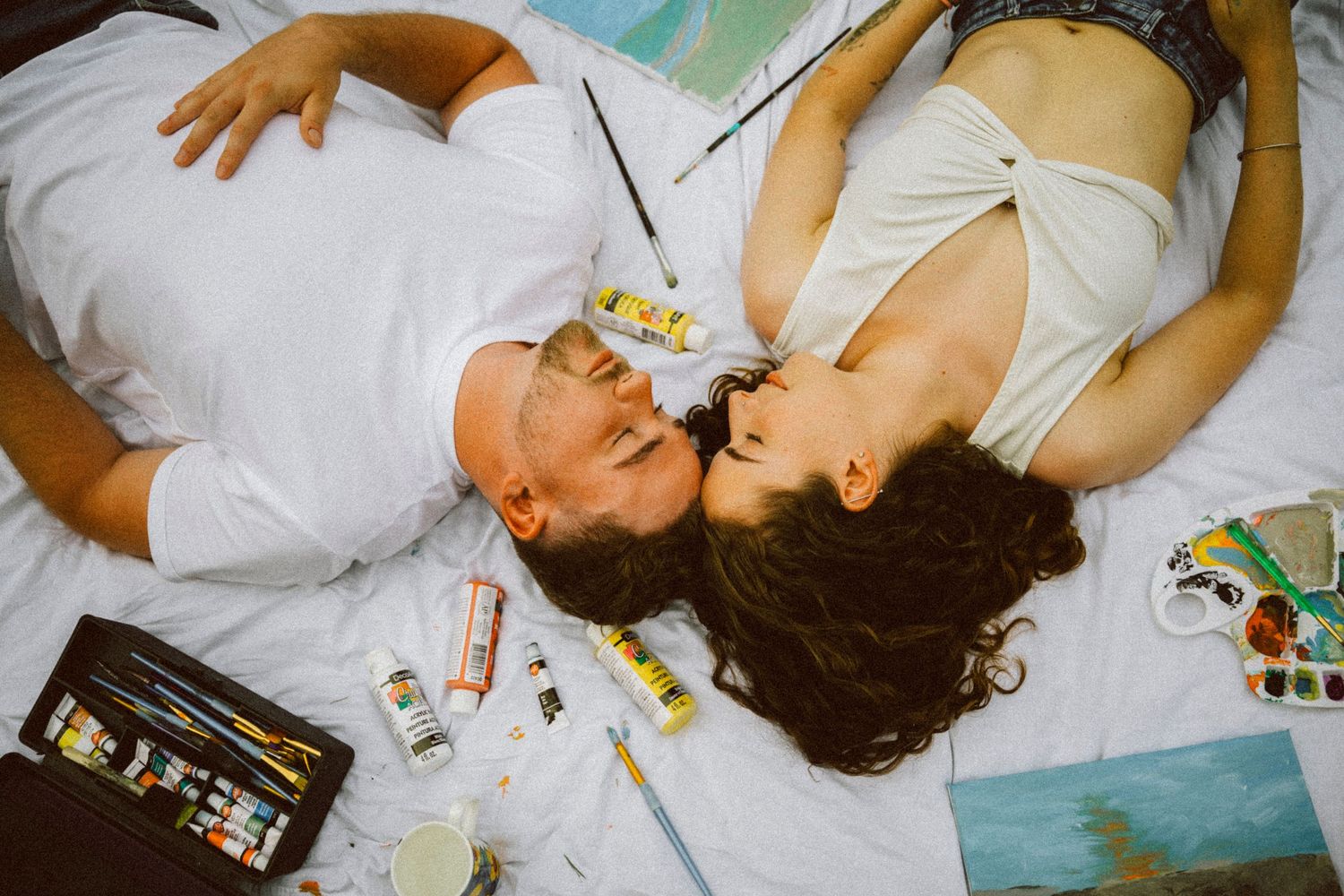 Overhead view of couple kissing while lying on blanket surrounded by paint tubes and brushes.