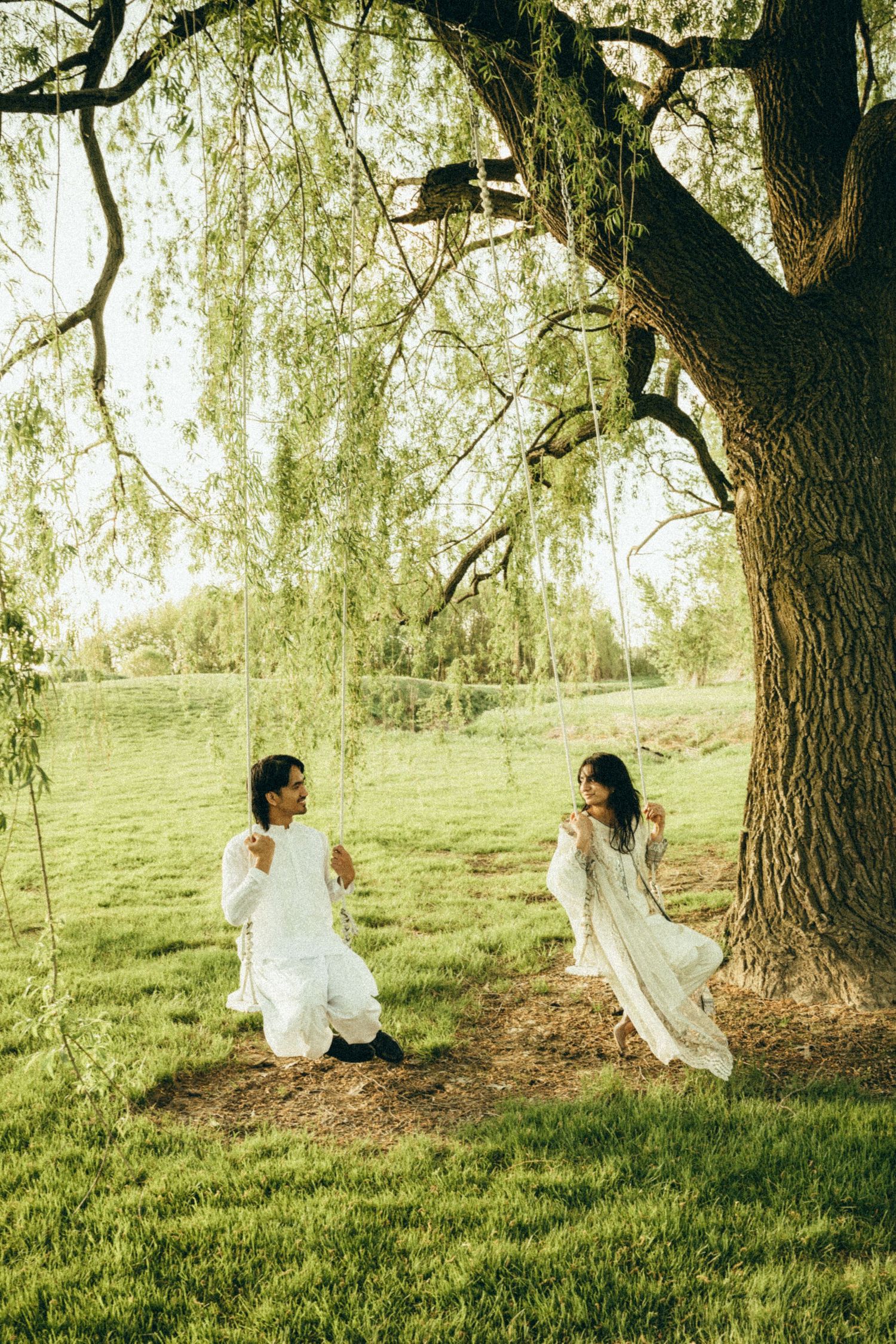 Couple in white attire sharing a moment on a swing under a willow tree.