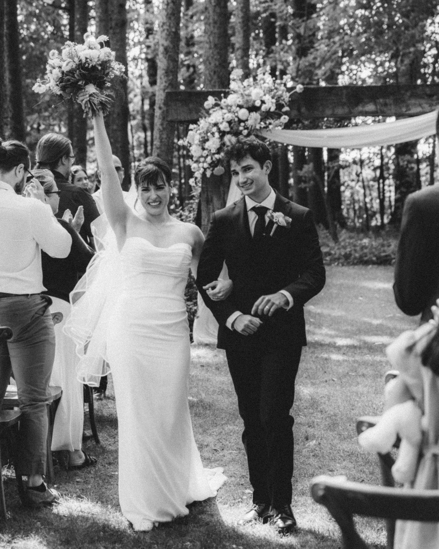Joyful newlyweds celebrate recessional in black and white photo, bride raising bouquet in woodland wedding ceremony.