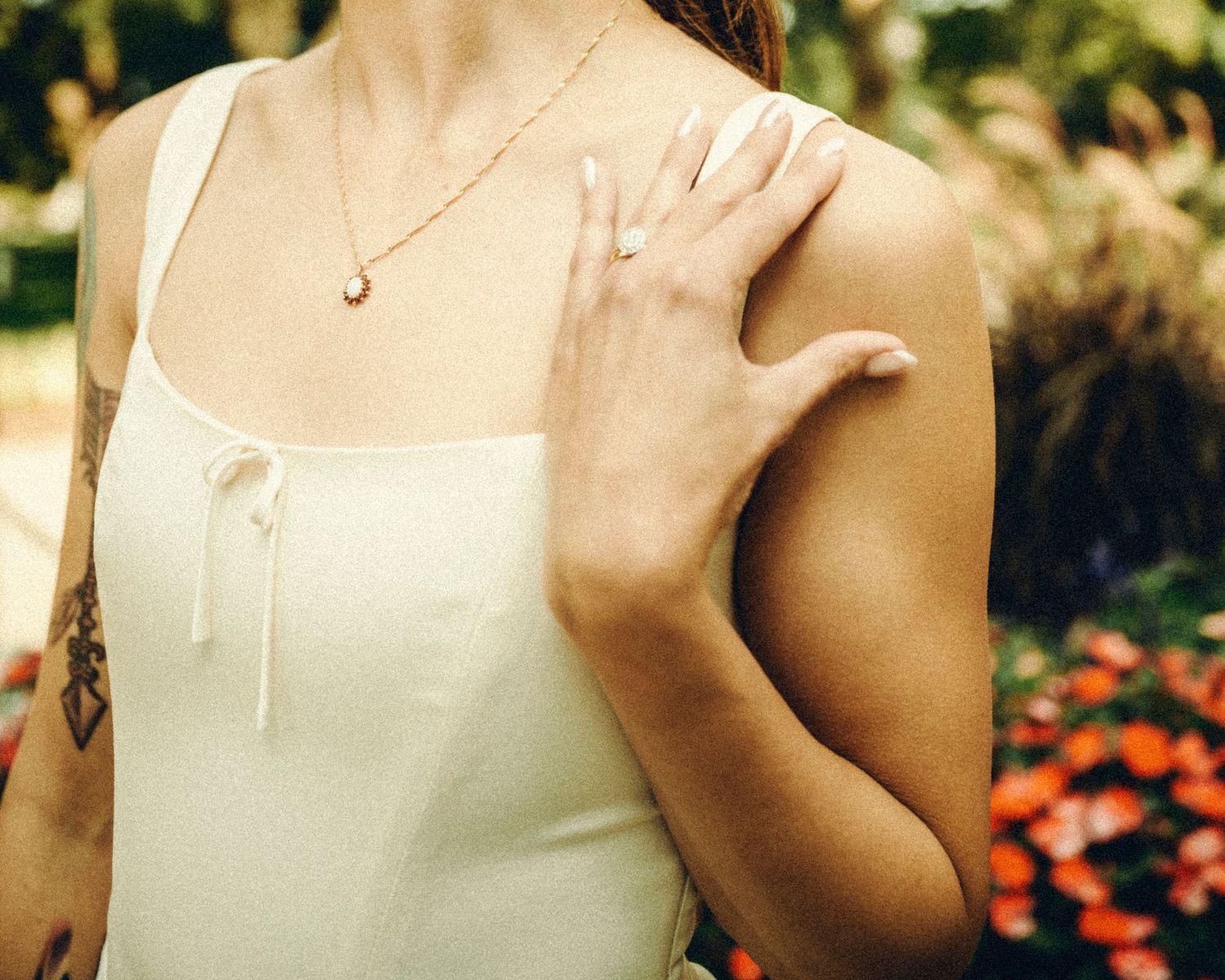 Bride in white dress with tattoo and gold necklace posing in garden setting.