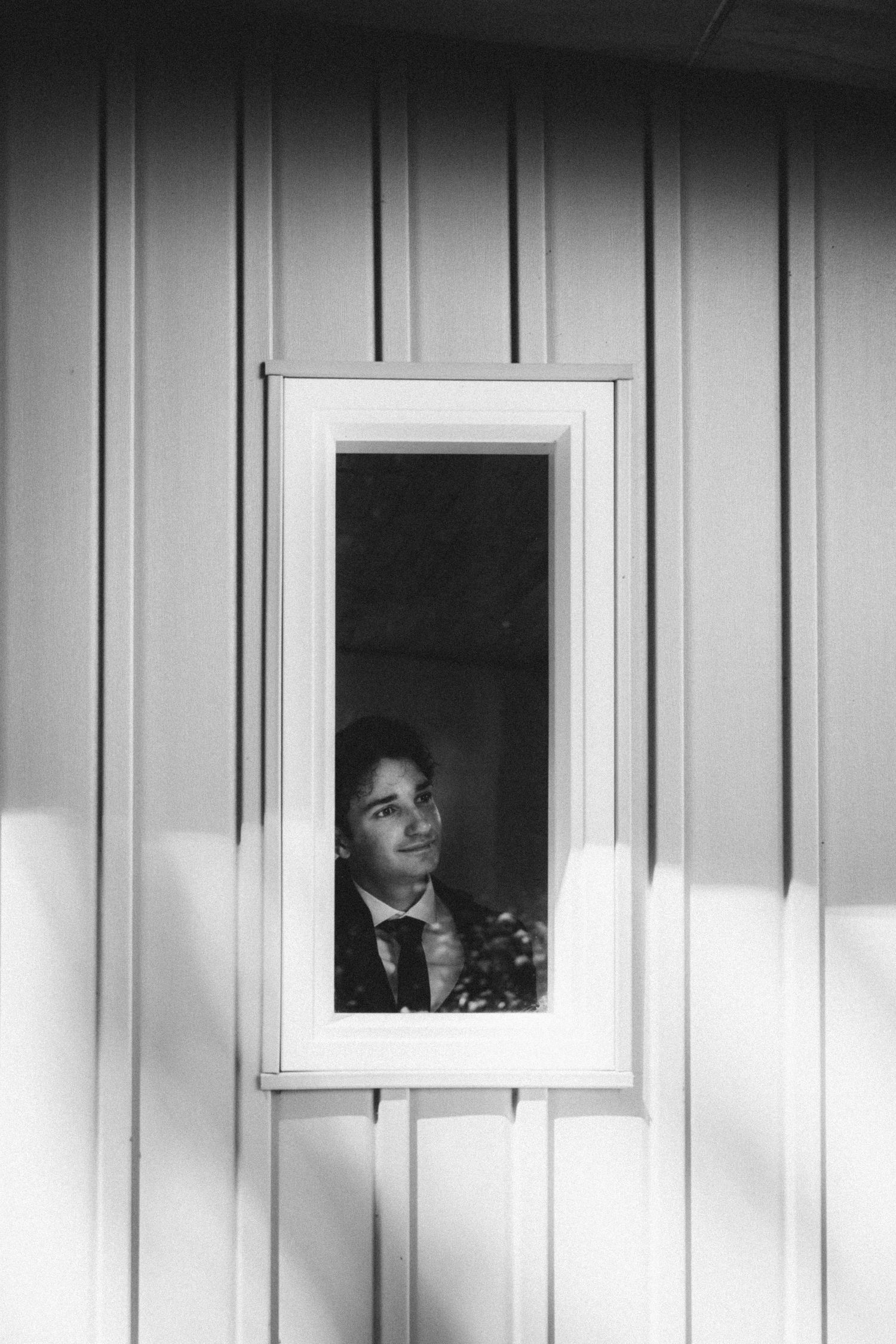 Groom in suit peers through a white-framed window of a wooden building in a black and white wedding photo.