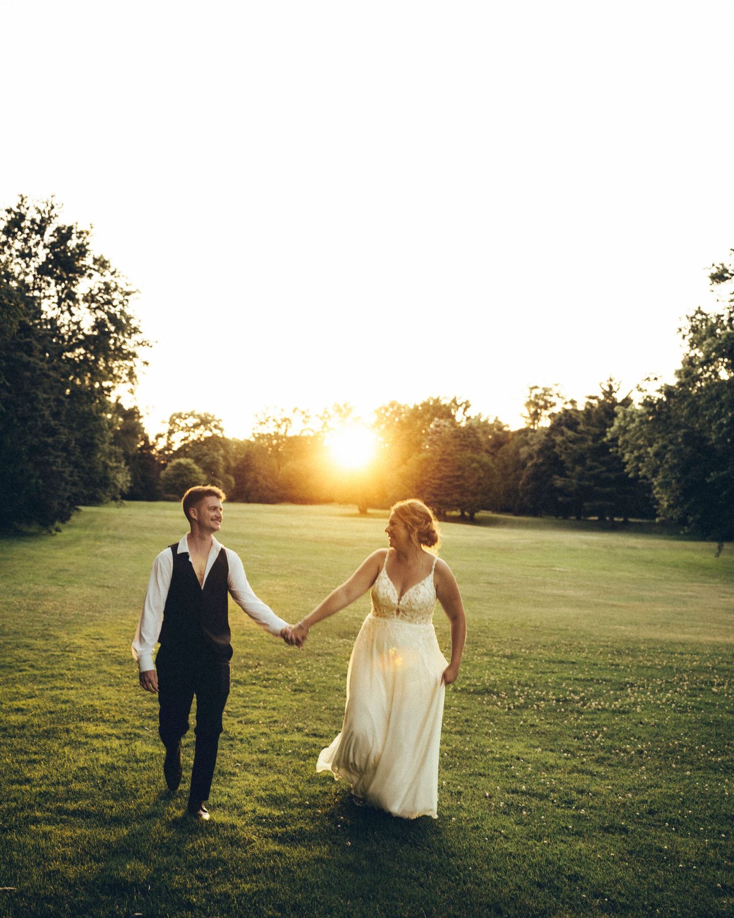 Bride in lace gown and groom in vest hold hands walking through sunlit green field.