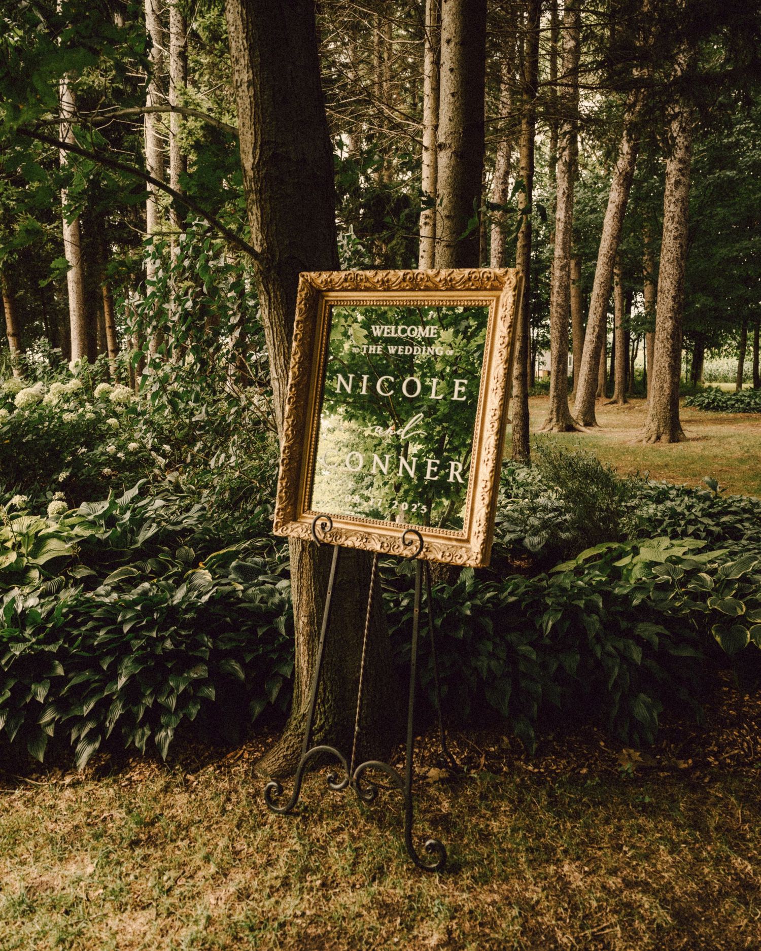 Ornate gold-framed wedding welcome sign for Nicole and Connor displayed on easel in wooded garden.