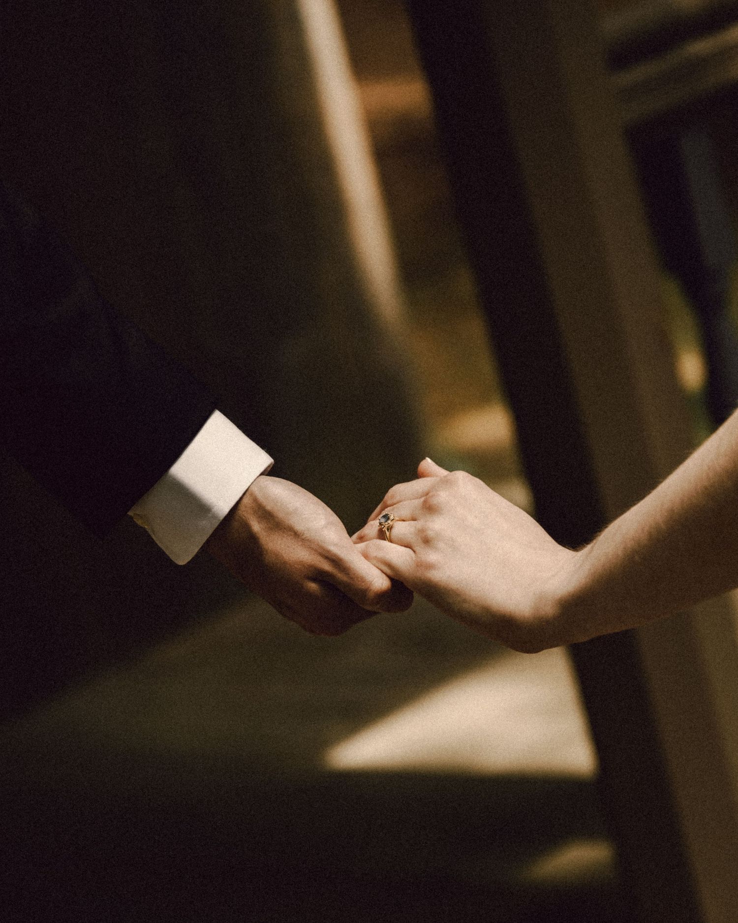 Close-up of bride and groom holding hands, showcasing wedding ring and groom's formal suit cufflink.