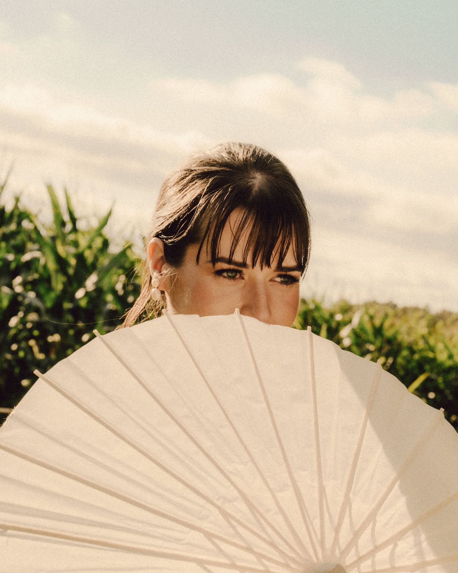 Close-up of bride with dark hair peeking over a white paper parasol with greenery background.