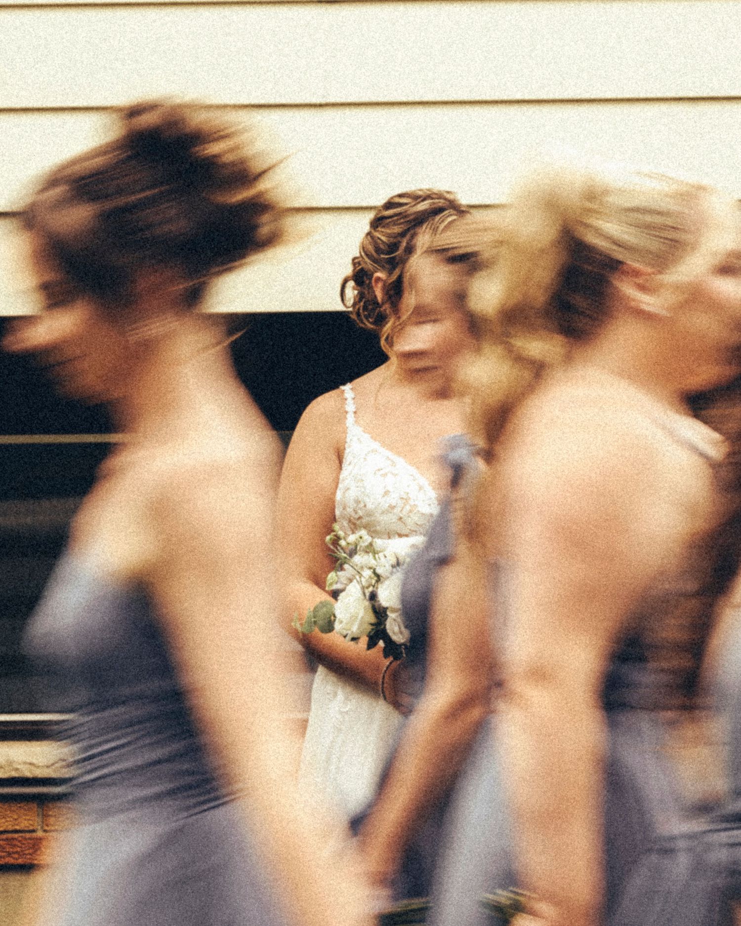 Bride in white lace gown holding bouquet surrounded by blurred bridesmaids in lavender dresses.