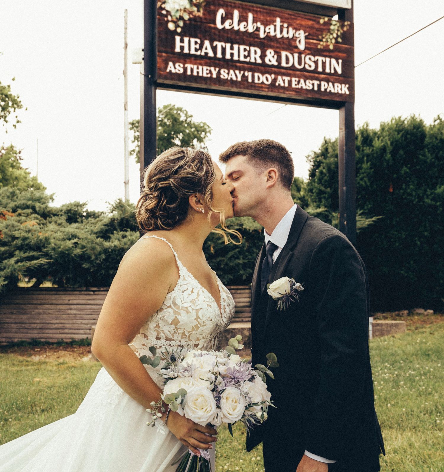 Bride and groom kiss in front of East Park celebrating Heather and Dustin wedding sign.