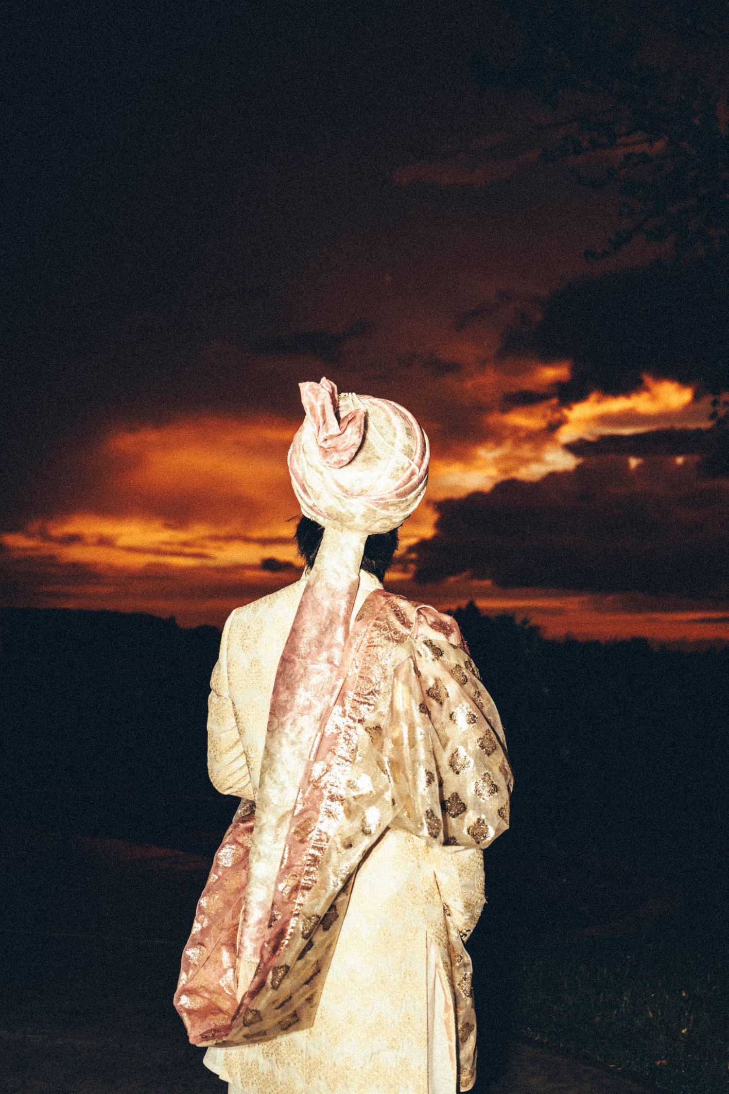South Asian groom in traditional ivory sherwani and turban stands silhouetted against a vivid orange sunset.
