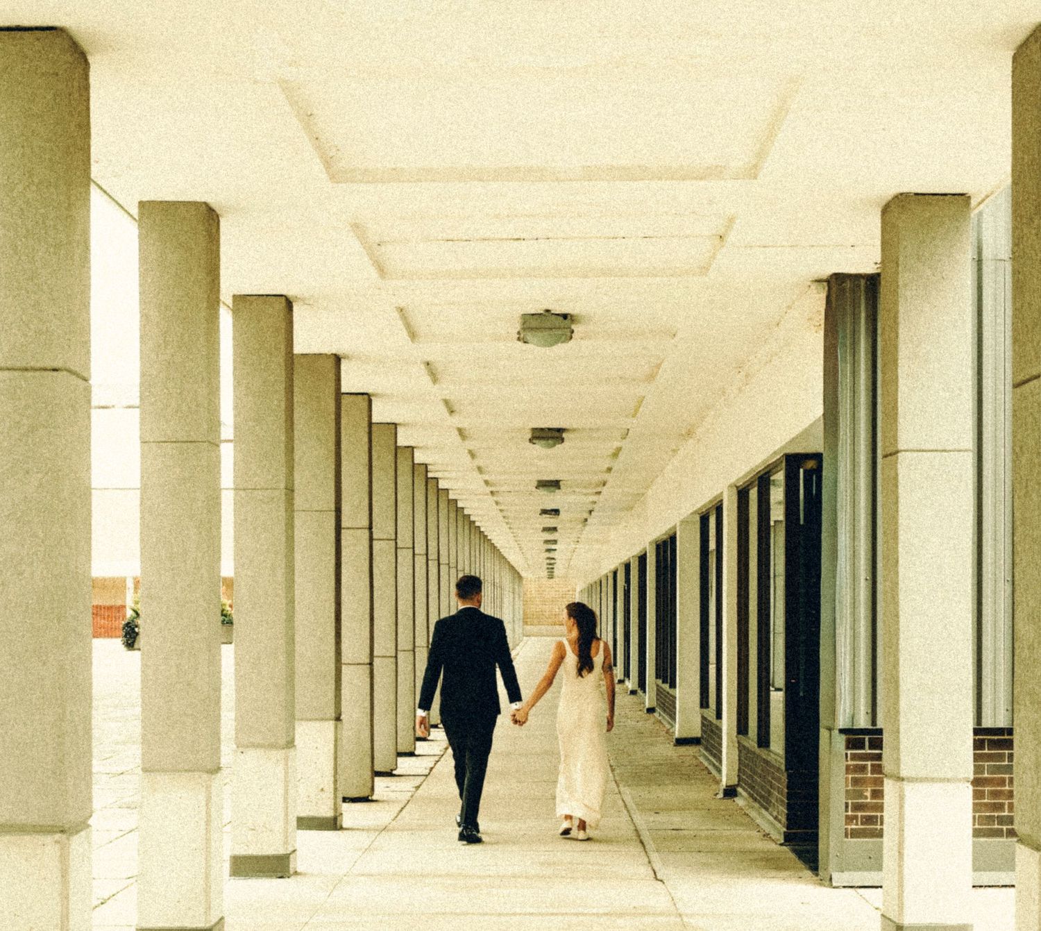 Newlyweds strolling hand in hand down a sunlit corridor lined with concrete columns.