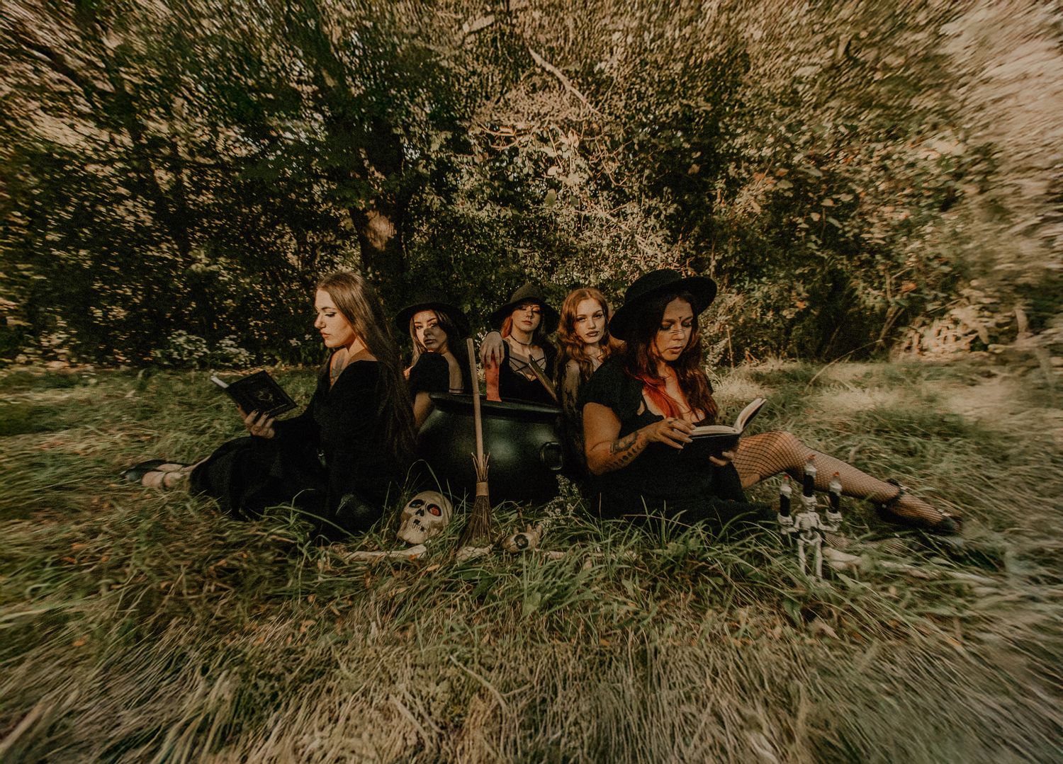 Four women dressed in black sitting in tall grass under a tree in a moody outdoor scene.