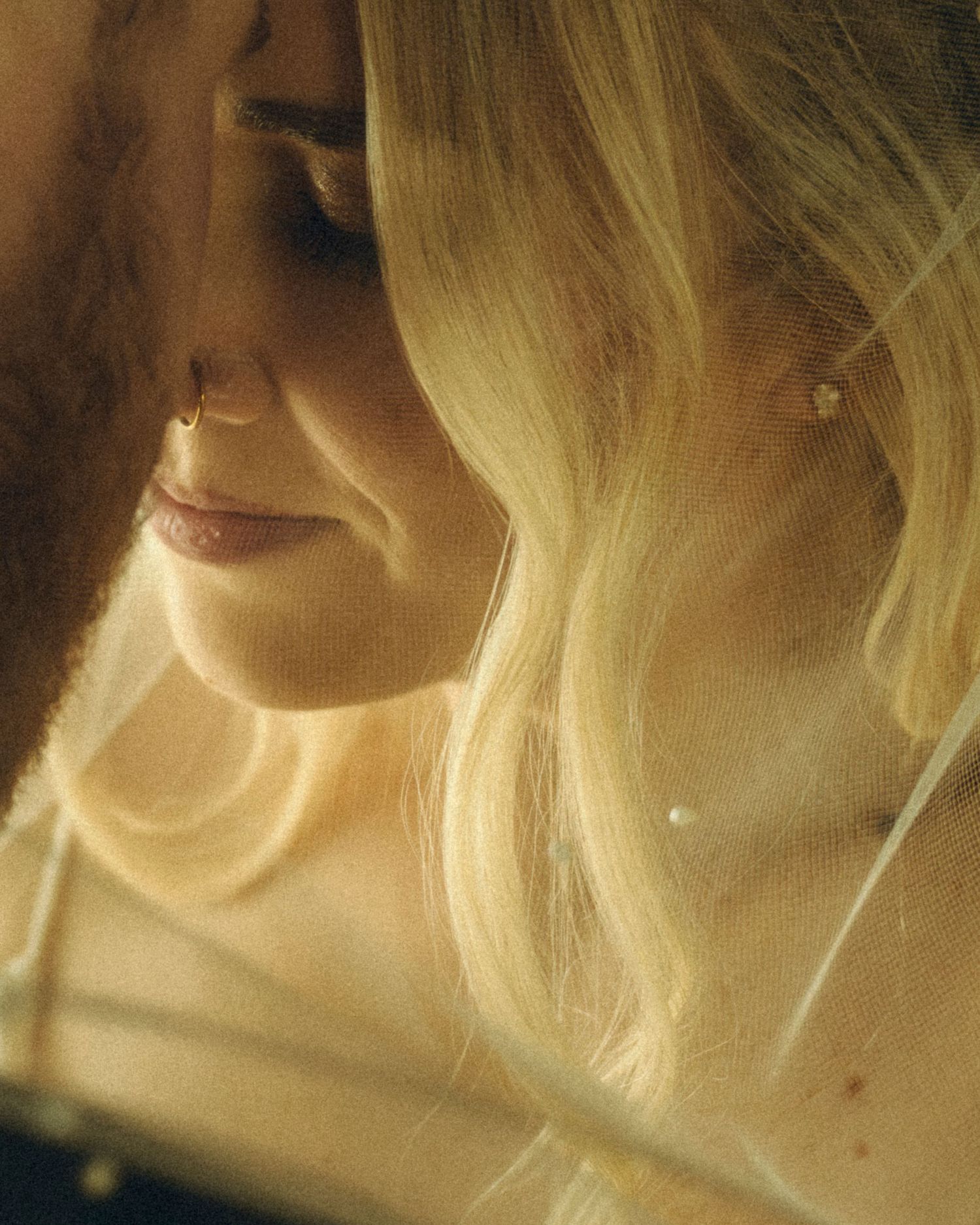 Close-up of smiling blonde bride wearing veil and gold nose ring in warm golden light.