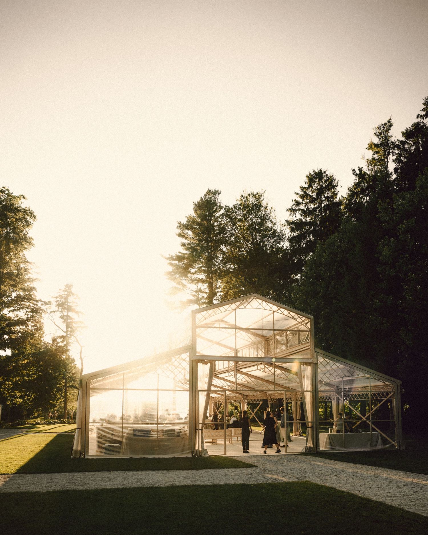 Glass wedding reception tent glowing at golden hour surrounded by trees.