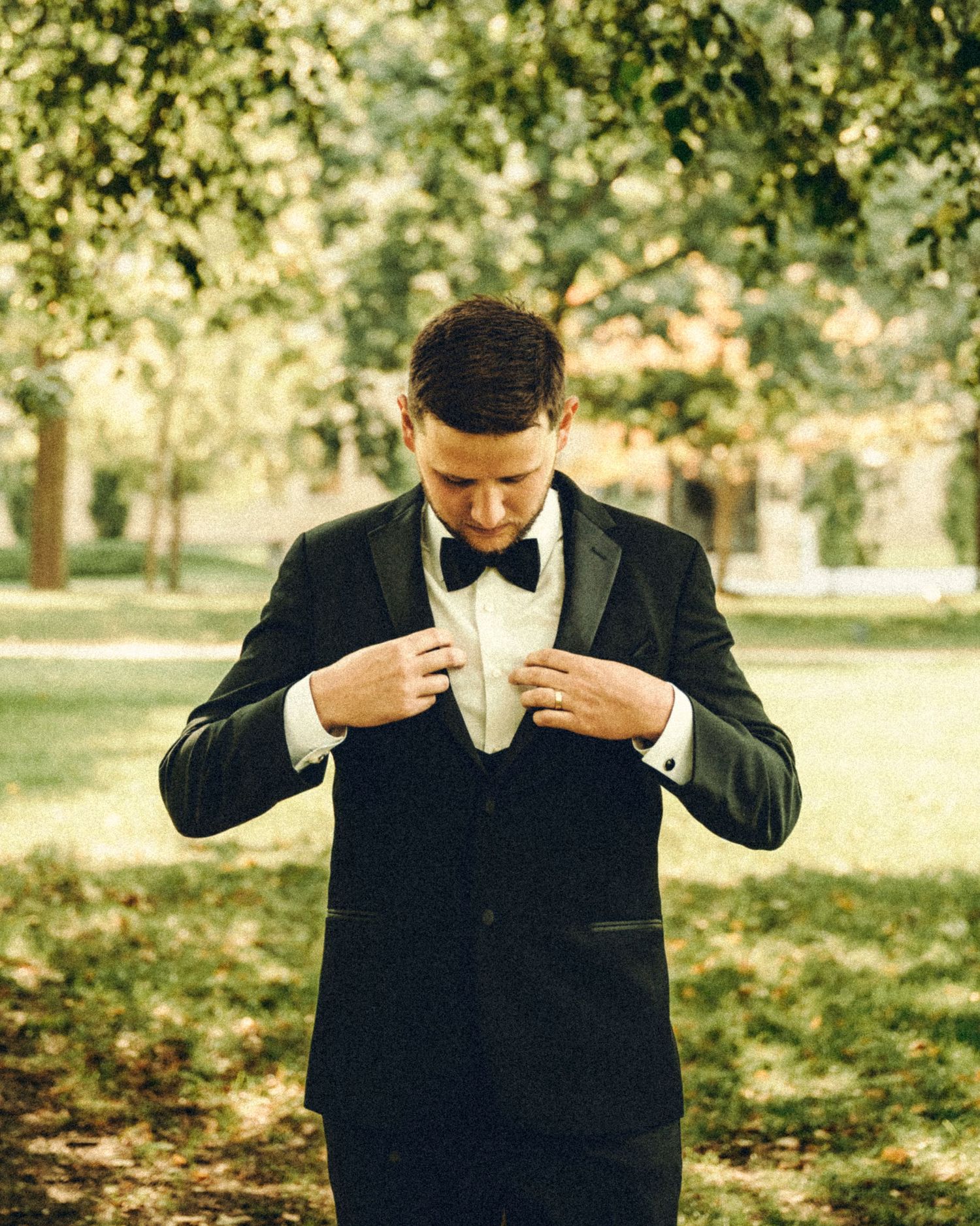 Dapper groom in black tuxedo adjusting jacket lapels while standing in a green park.