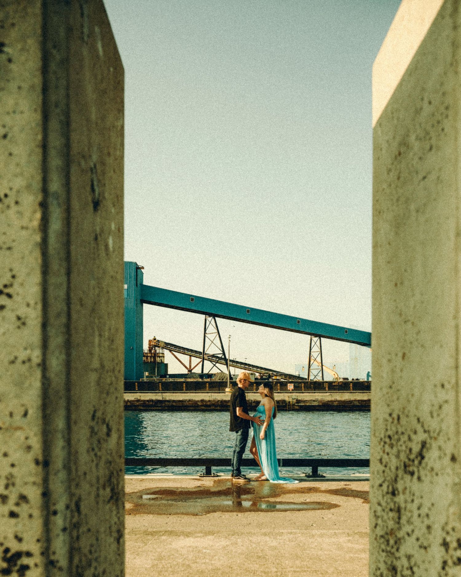 Couple in teal gown kissing by waterfront framed between concrete pillars near bridge.