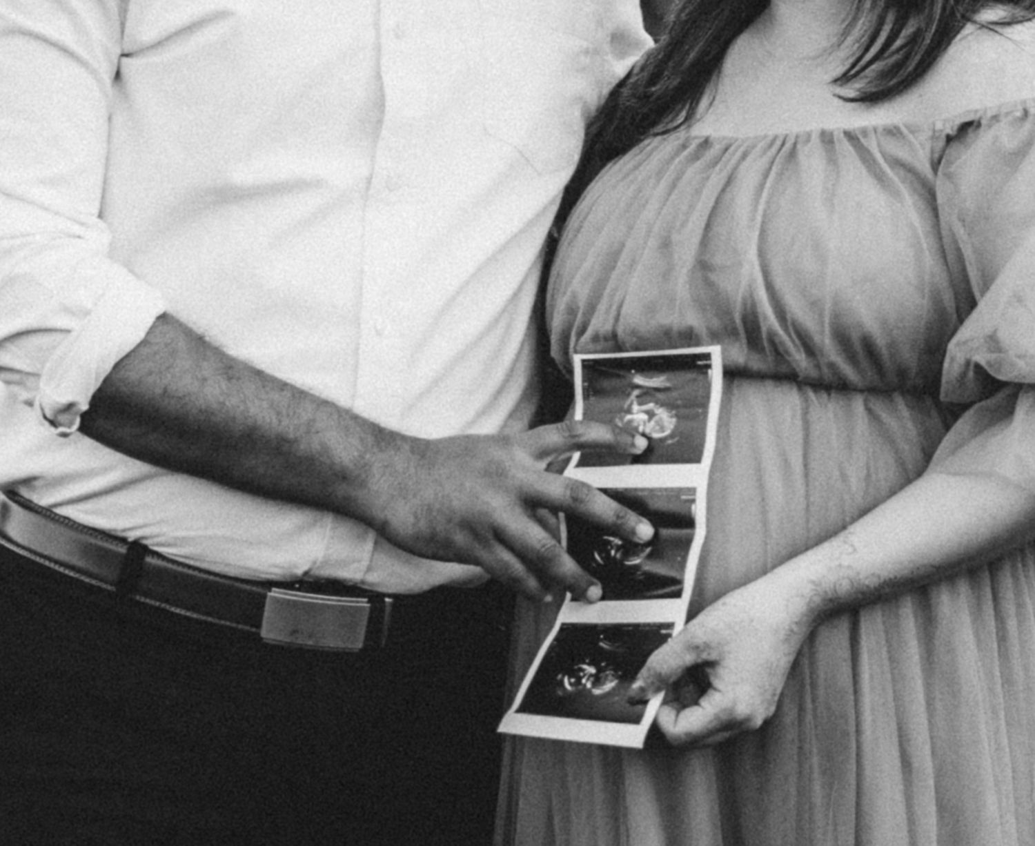 Black and white photo of pregnant couple holding ultrasound photos against a maternity dress bump.