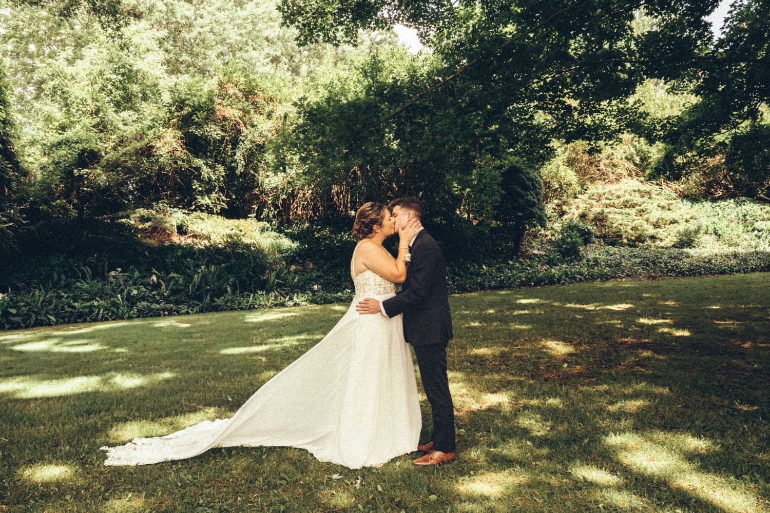 Bride and groom sharing a kiss on a sunlit green lawn surrounded by lush trees.